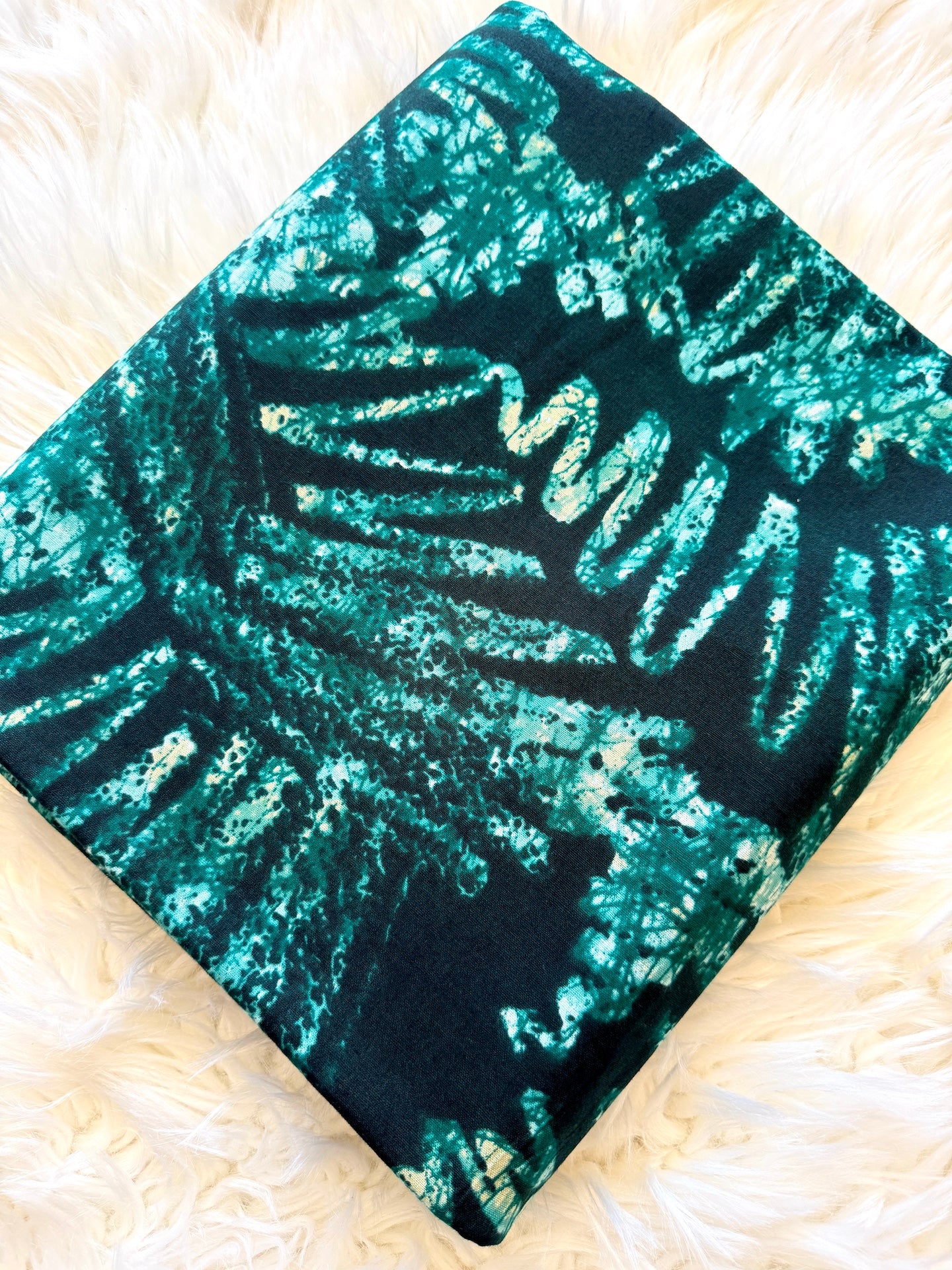 A square fabric item, likely a small blanket or throw, featuring a glittery teal and black tropical leaf pattern, laid flat on a white faux fur surface.