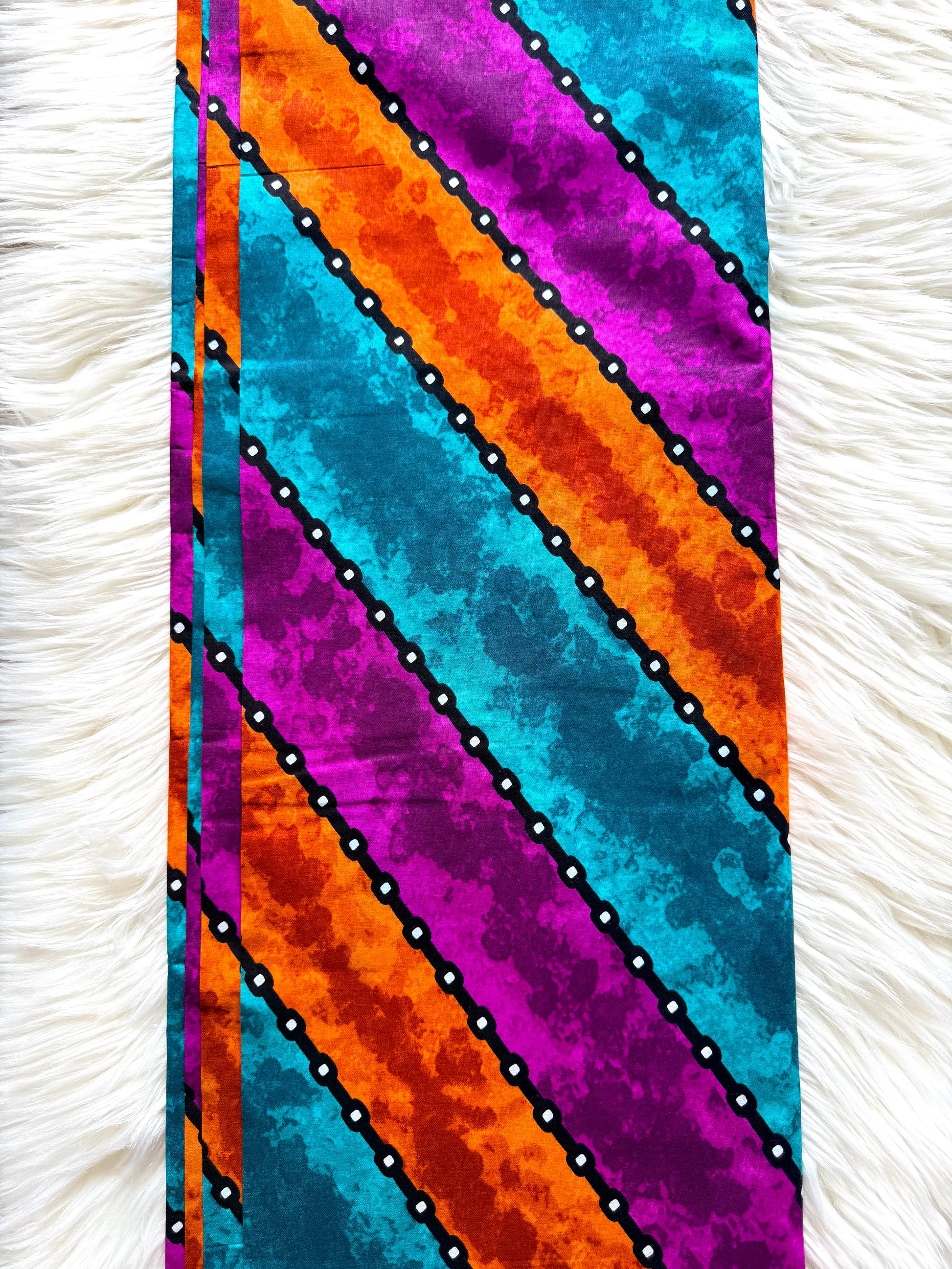 A vertical fabric panel or scarf with a bold diagonal striped pattern in turquoise, orange, magenta, and purple, separated by black dotted lines, laid flat on a white faux fur surface.