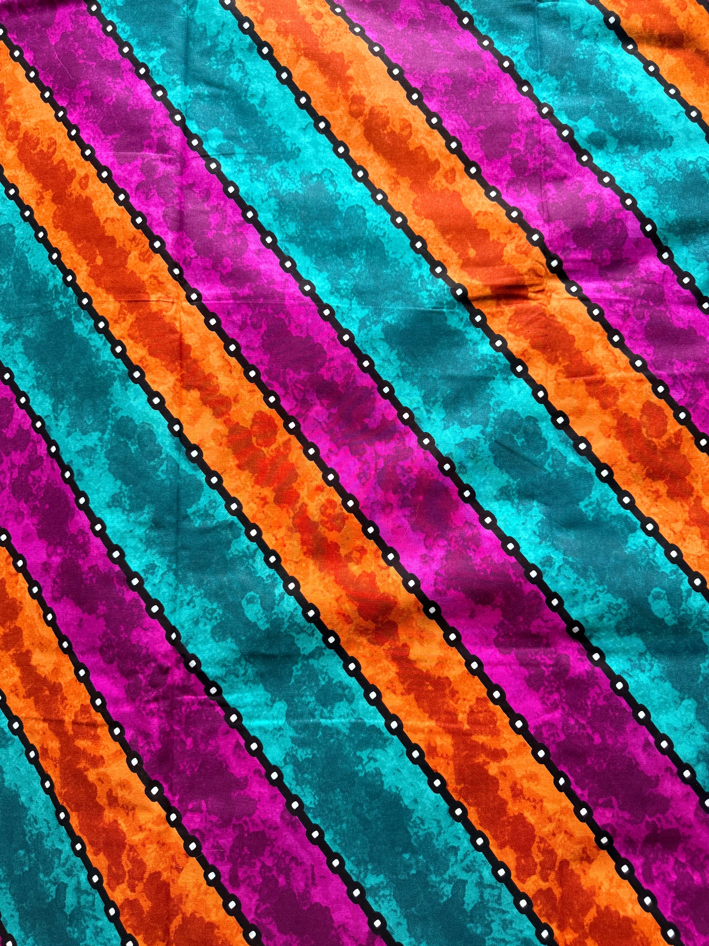 A close-up of a colorful striped fabric pattern with diagonal bands of turquoise, orange, magenta, and purple separated by black dotted lines with white dots.