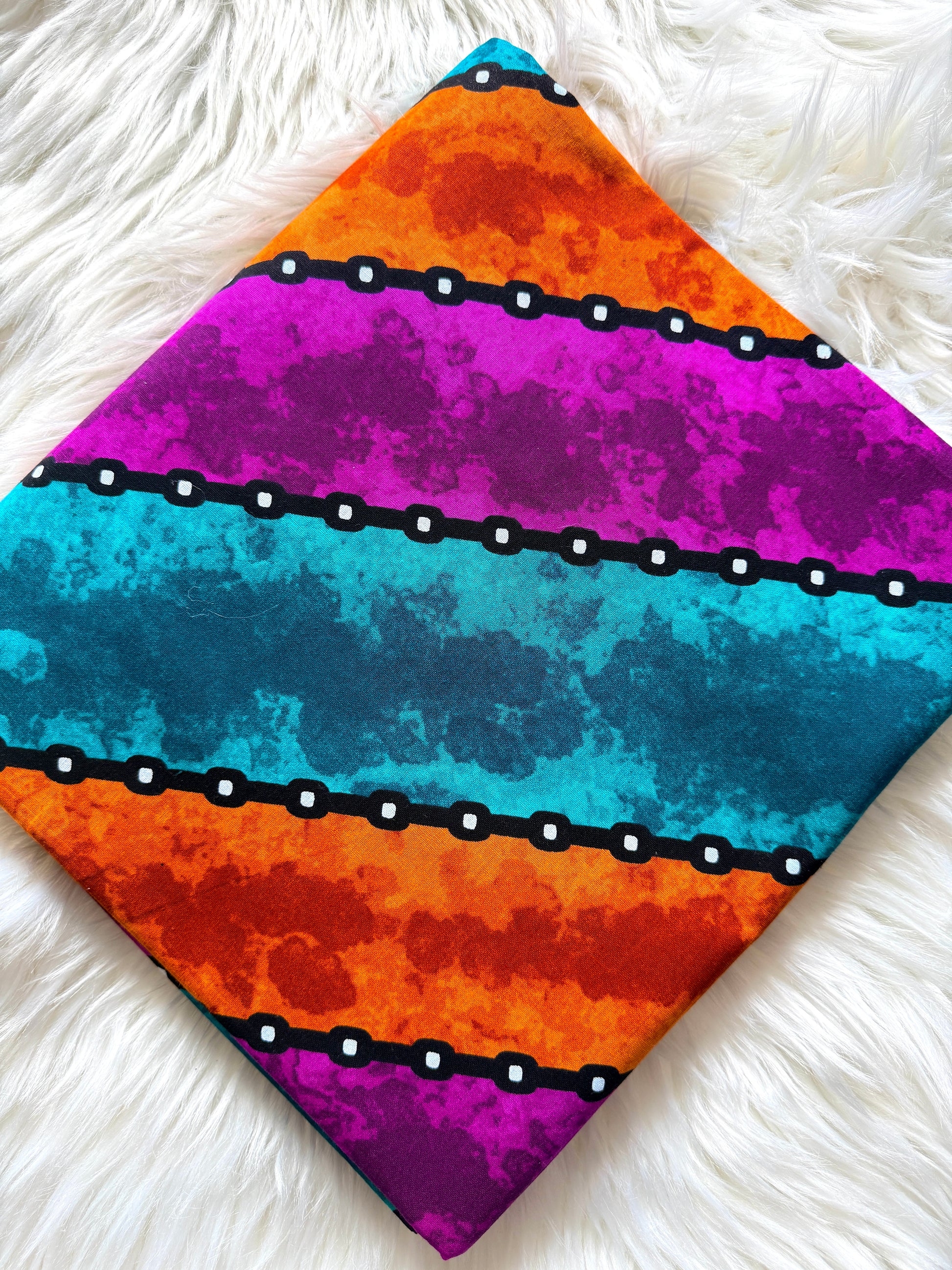A square fabric item, likely a napkin or small scarf, featuring a vibrant horizontal striped pattern with orange, magenta, and teal watercolor textures, separated by black bands with white dots, laid flat on a white faux fur surface.