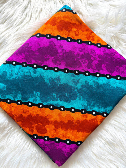 A square fabric item, likely a napkin or small scarf, featuring a vibrant horizontal striped pattern with orange, magenta, and teal watercolor textures, separated by black bands with white dots, laid flat on a white faux fur surface.
