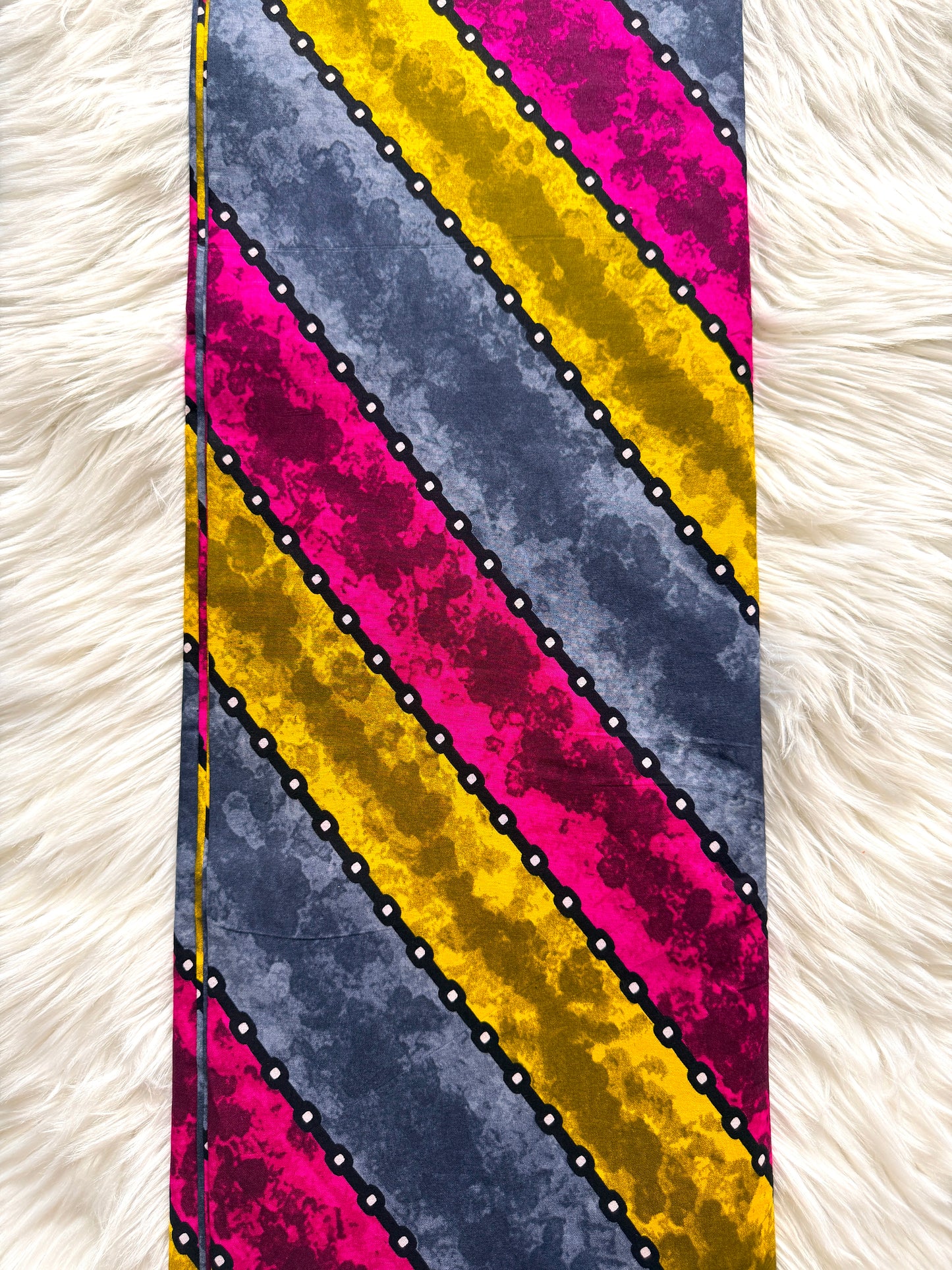 A close-up of a striped fabric pattern with diagonal bands of navy blue, bright pink, and mustard yellow, separated by black dotted lines with small white dots, laid flat on a white faux fur background.