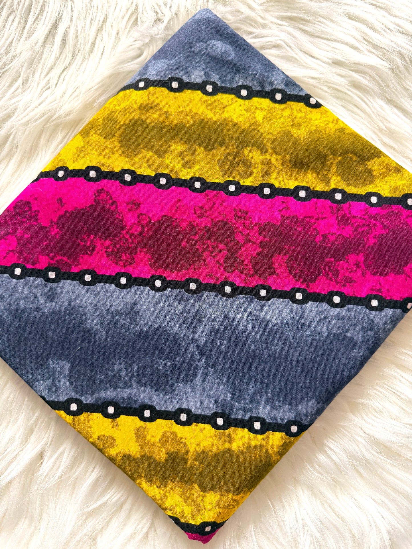 A square fabric accessory, likely a pocket square or small scarf, featuring a bold horizontal striped pattern with yellow, magenta, and slate blue sections separated by black chains with white dots, displayed on a white faux fur background.