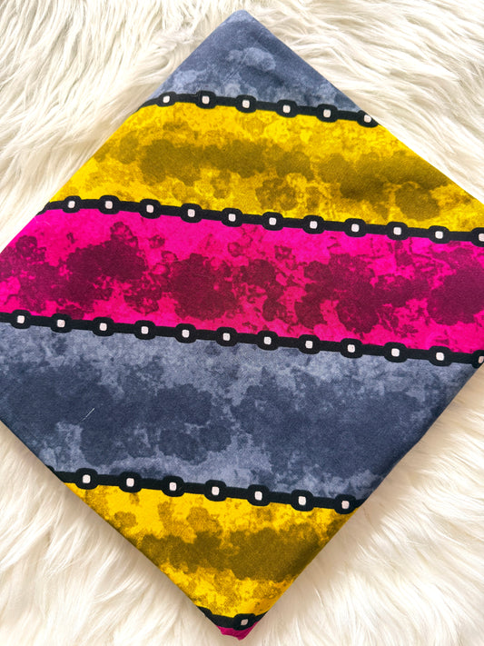 A square fabric accessory, likely a pocket square or small scarf, featuring a bold horizontal striped pattern with yellow, magenta, and slate blue sections separated by black chains with white dots, displayed on a white faux fur background.