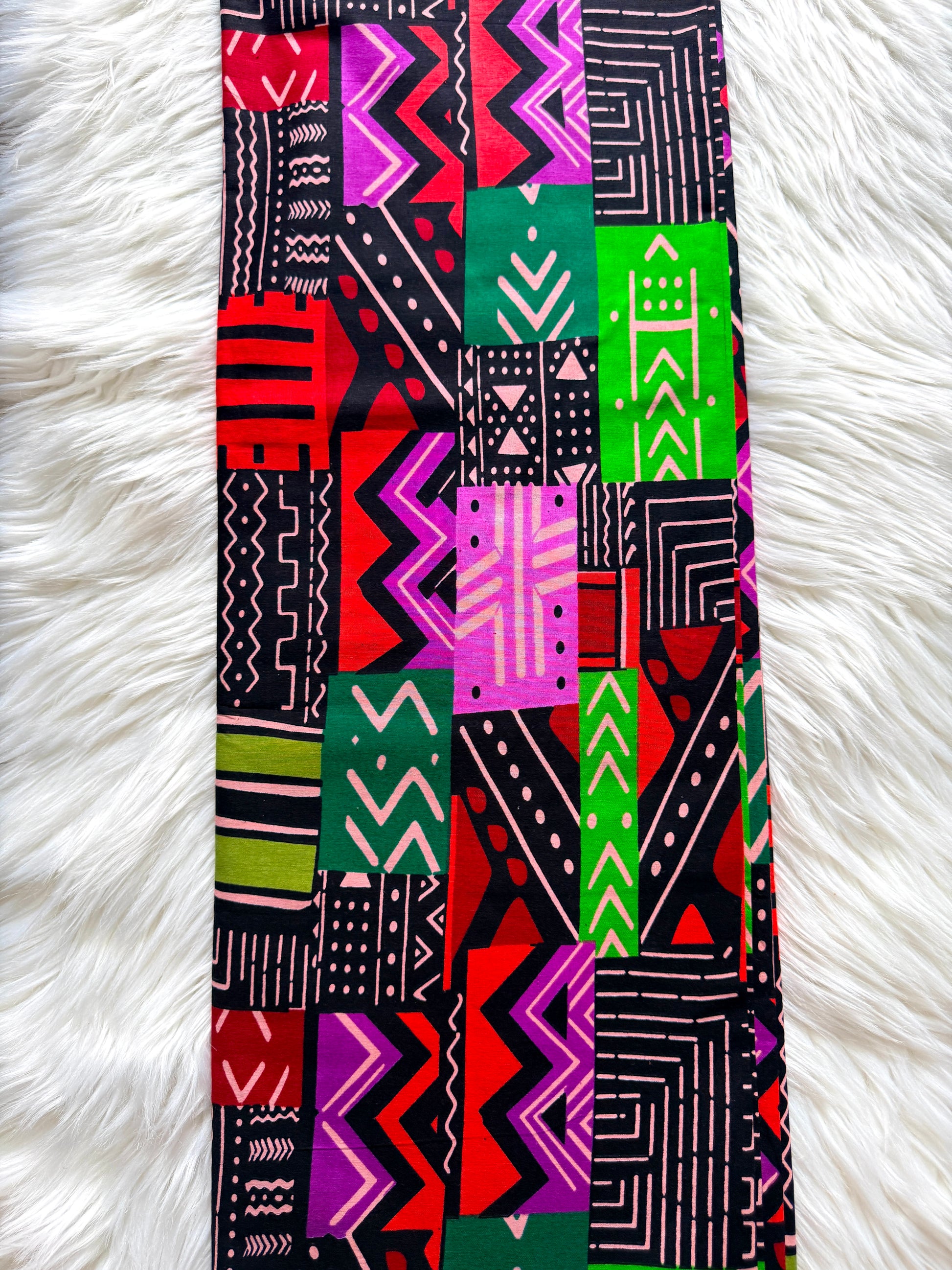 A long rectangular scarf with a vibrant multicolor geometric patchwork pattern, laid flat on a white faux fur surface.