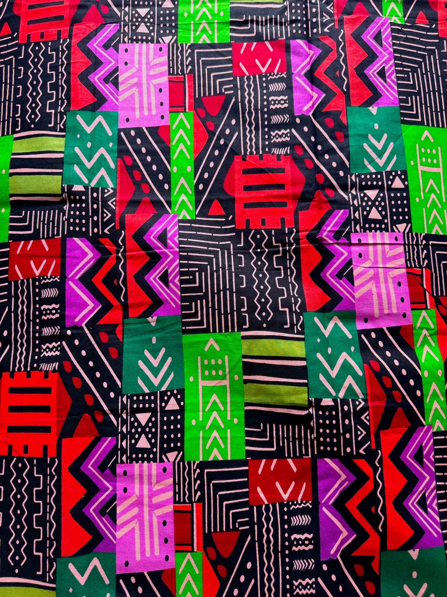 A close-up of a colorful patchwork fabric featuring various African wax print patterns in red, green, purple, black, and white.