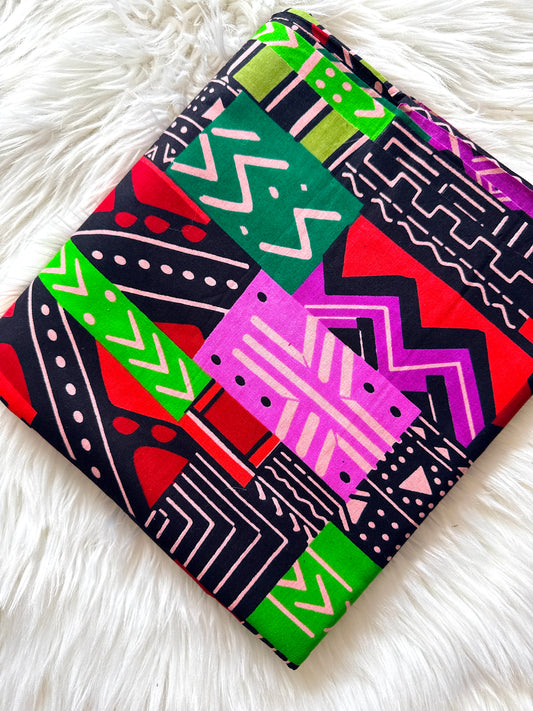 A folded square fabric item, likely a napkin, small towel, or scarf, featuring a vibrant geometric patchwork print with pink, green, red, and black tribal patterns, displayed on a white faux fur surface.