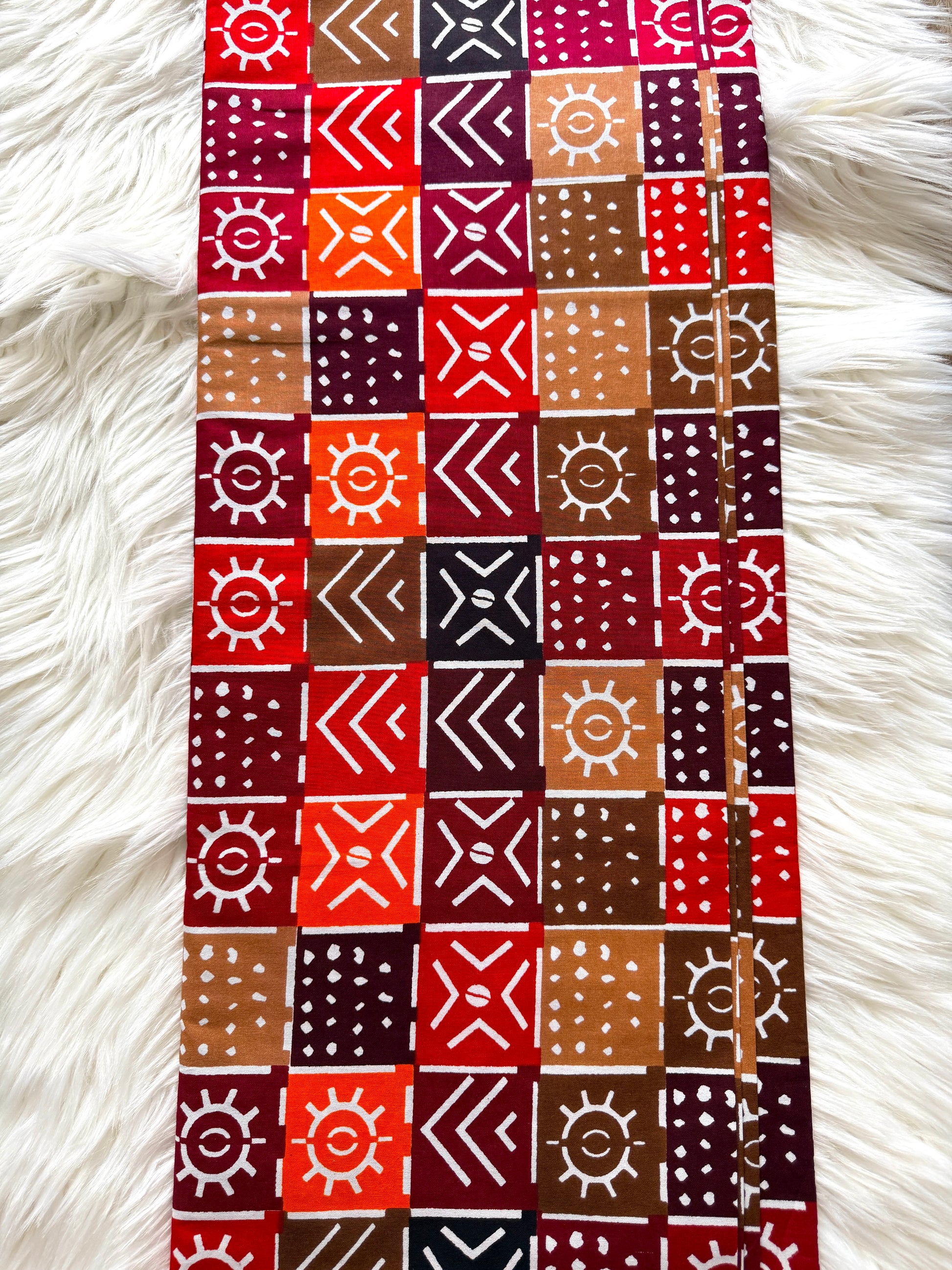 A long rectangular textile laid flat on a white faux-fur surface, featuring a repeating patchwork pattern of red, orange, brown, burgundy, black, and beige geometric and sun-like motifs.