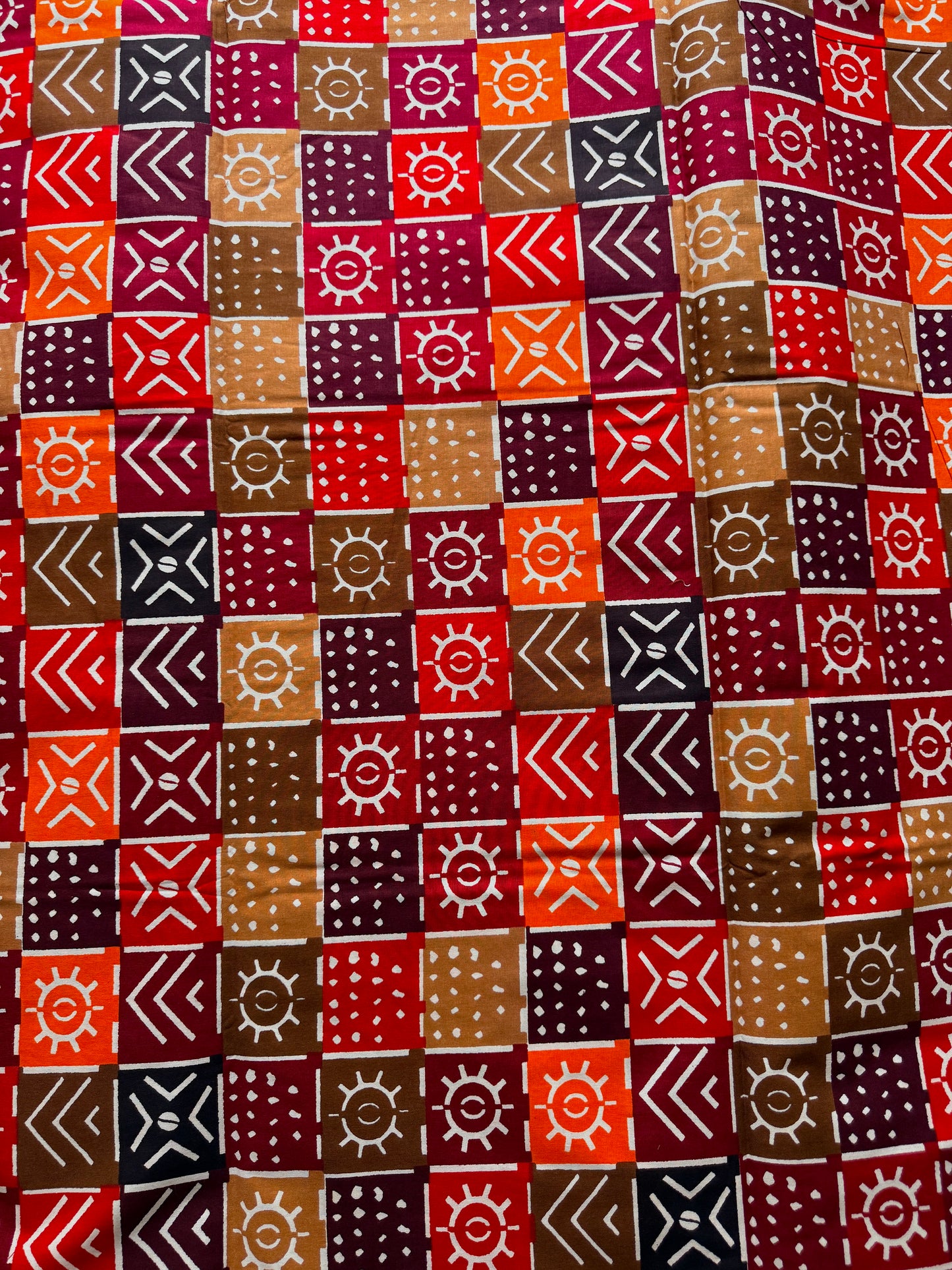 A close-up of a patterned textile featuring a grid of square blocks in red, orange, brown, beige, and dark navy, each decorated with white geometric and sun-like motifs.