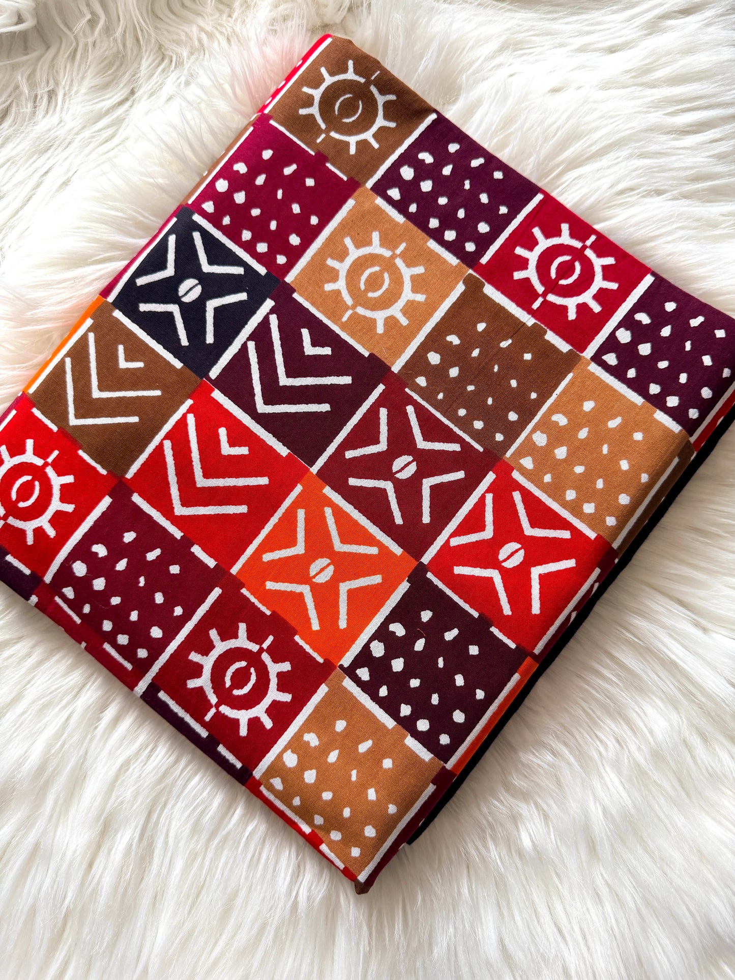 A square fabric item, likely a scarf or bandana, featuring a patchwork design of red, orange, brown, and dark purple squares with white geometric and sun-like patterns, laid flat on a white faux fur surface.