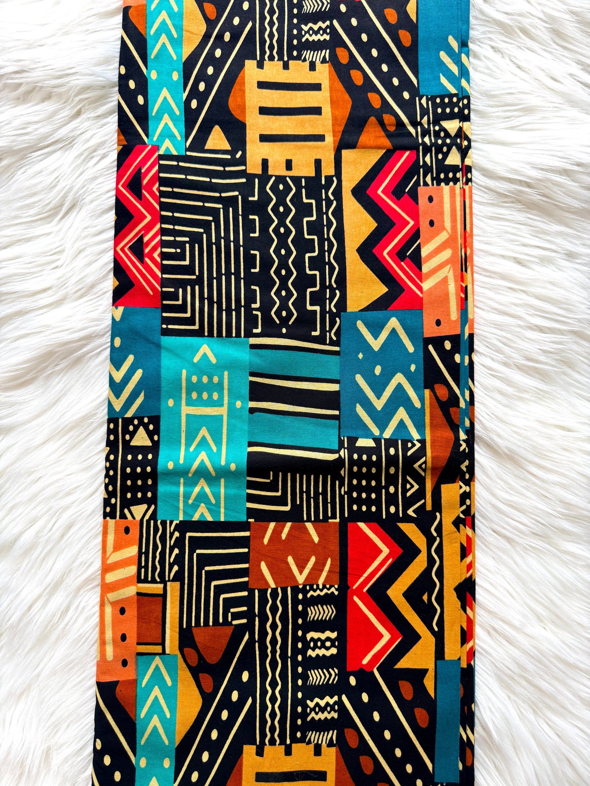 A vertical rectangular fabric item, likely a scarf or narrow wrap, featuring a vibrant patchwork of African tribal patterns in turquoise, orange, red, black, and cream, laid flat on a white faux fur surface.