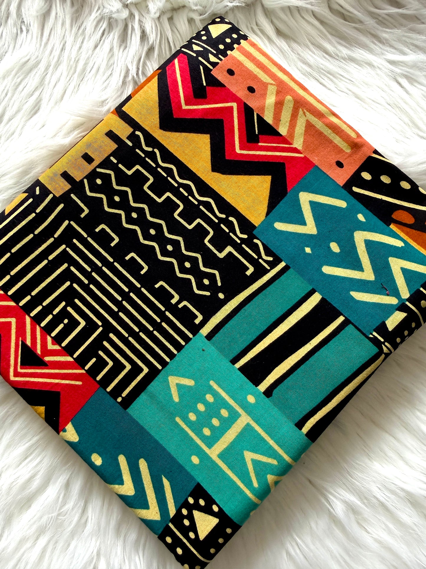 A square fabric item, likely a pillow cover or small blanket, featuring a vibrant geometric patchwork design with tribal-inspired patterns in black, teal, orange, red, and yellow, laid flat on a white faux fur surface.
