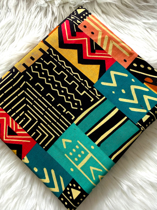 A square fabric item, likely a pillow cover or small blanket, featuring a vibrant geometric patchwork design with tribal-inspired patterns in black, teal, orange, red, and yellow, laid flat on a white faux fur surface.
