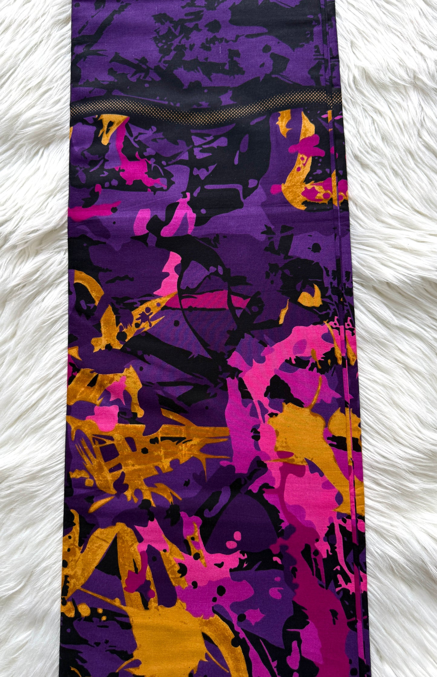 A close-up of a fabric product, likely leggings or a skirt, featuring an abstract paint-splatter pattern in purple, magenta, black, and mustard yellow, laid flat on a white faux fur surface.