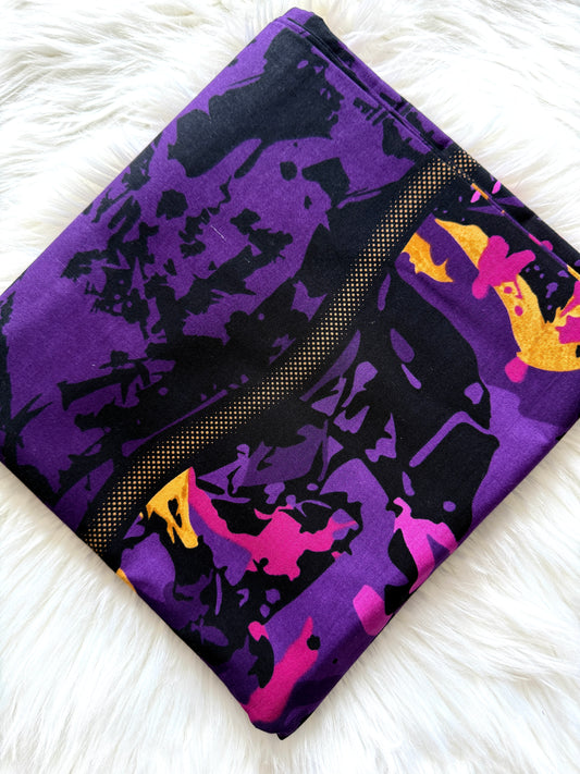 A folded fabric pouch or clutch with an abstract paint-splatter pattern in purple, black, pink, and yellow, resting on a white faux fur surface.