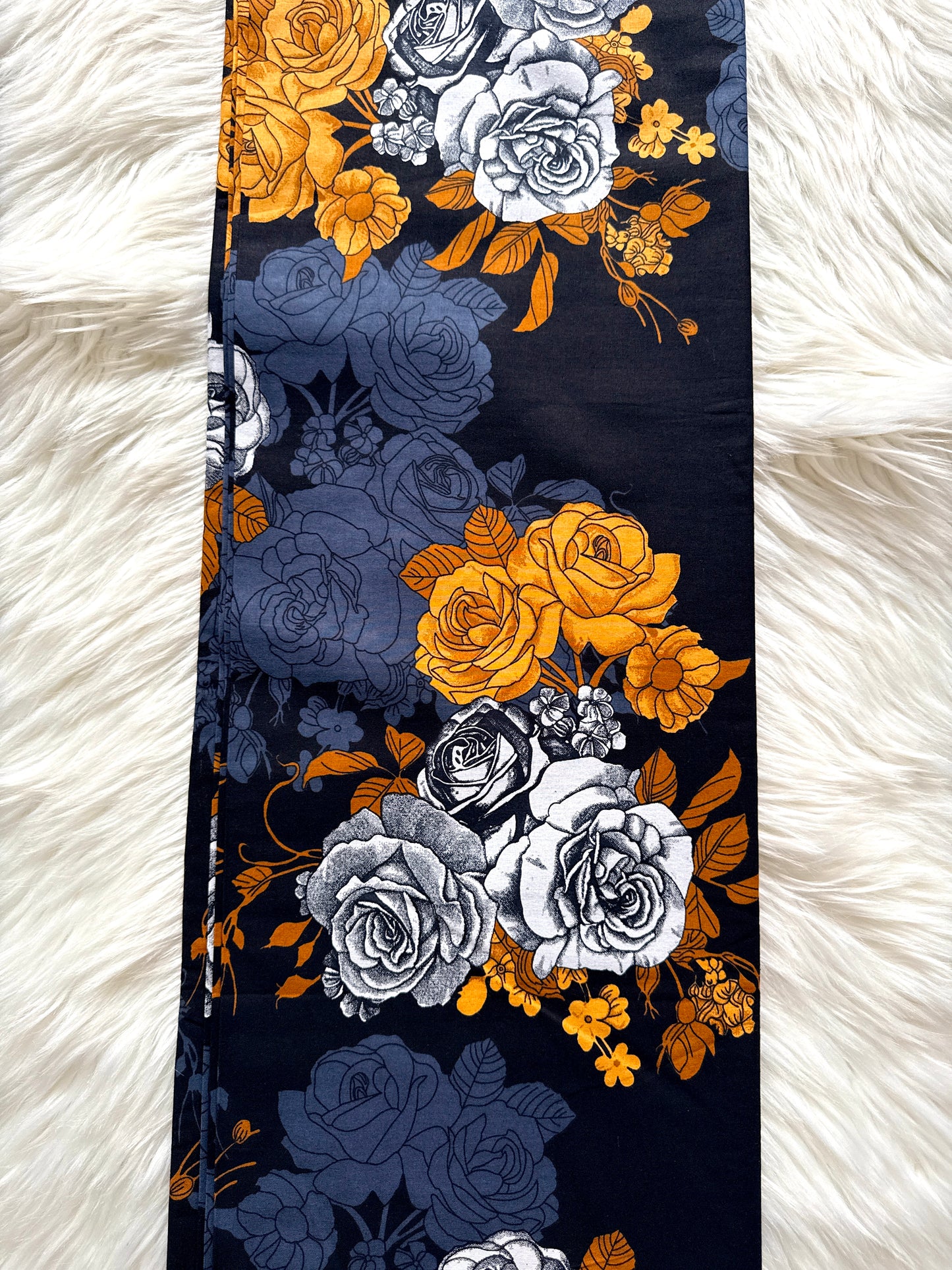 A long rectangular scarf laid flat on a white faux fur surface, featuring a floral rose pattern in navy blue, mustard yellow, and light gray.
