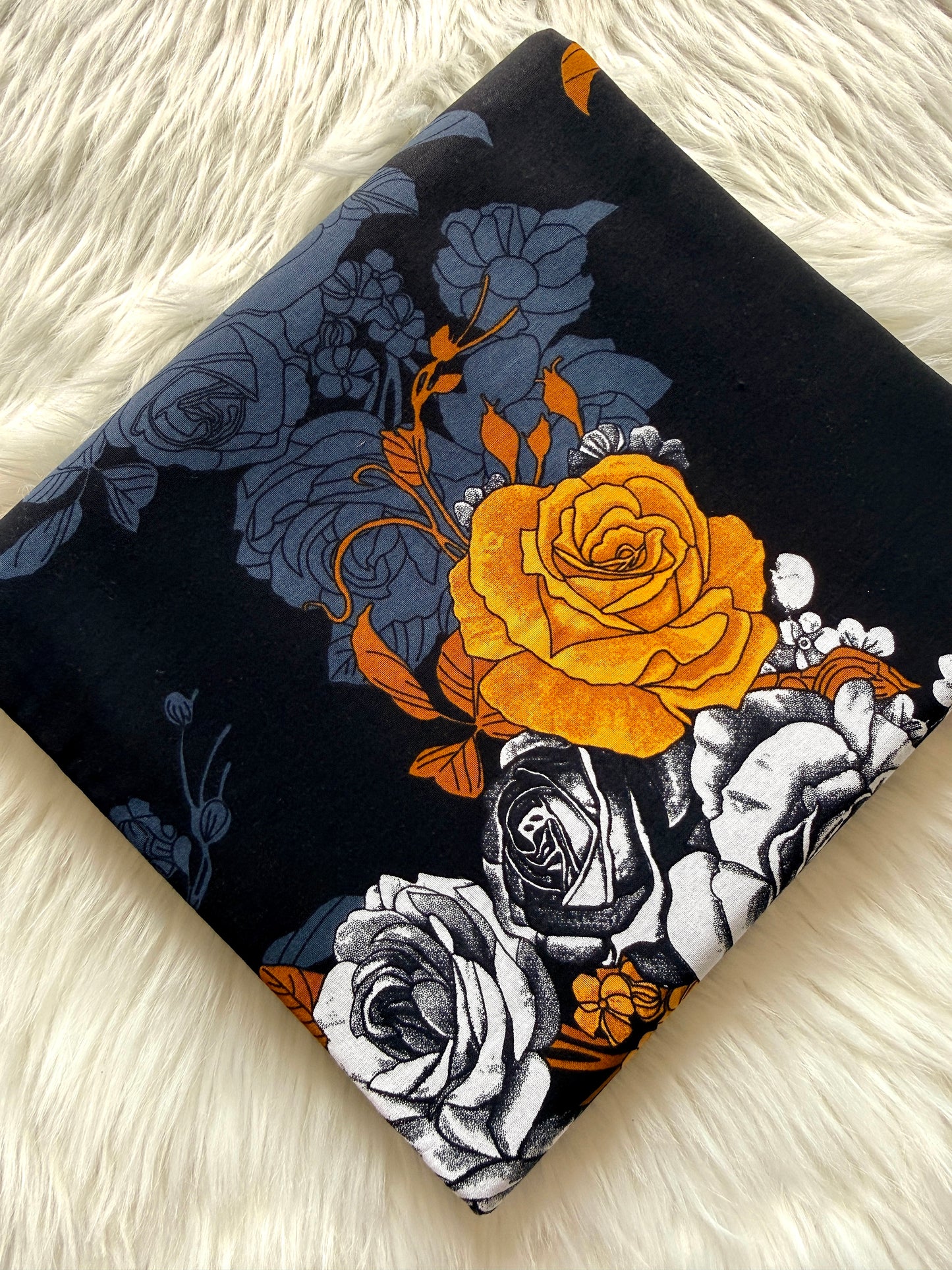 A square fabric item, likely a pillow cover or scarf, featuring a floral print with a prominent orange rose, white and gray roses, and blue-gray roses on a black background, styled on a white faux fur surface.