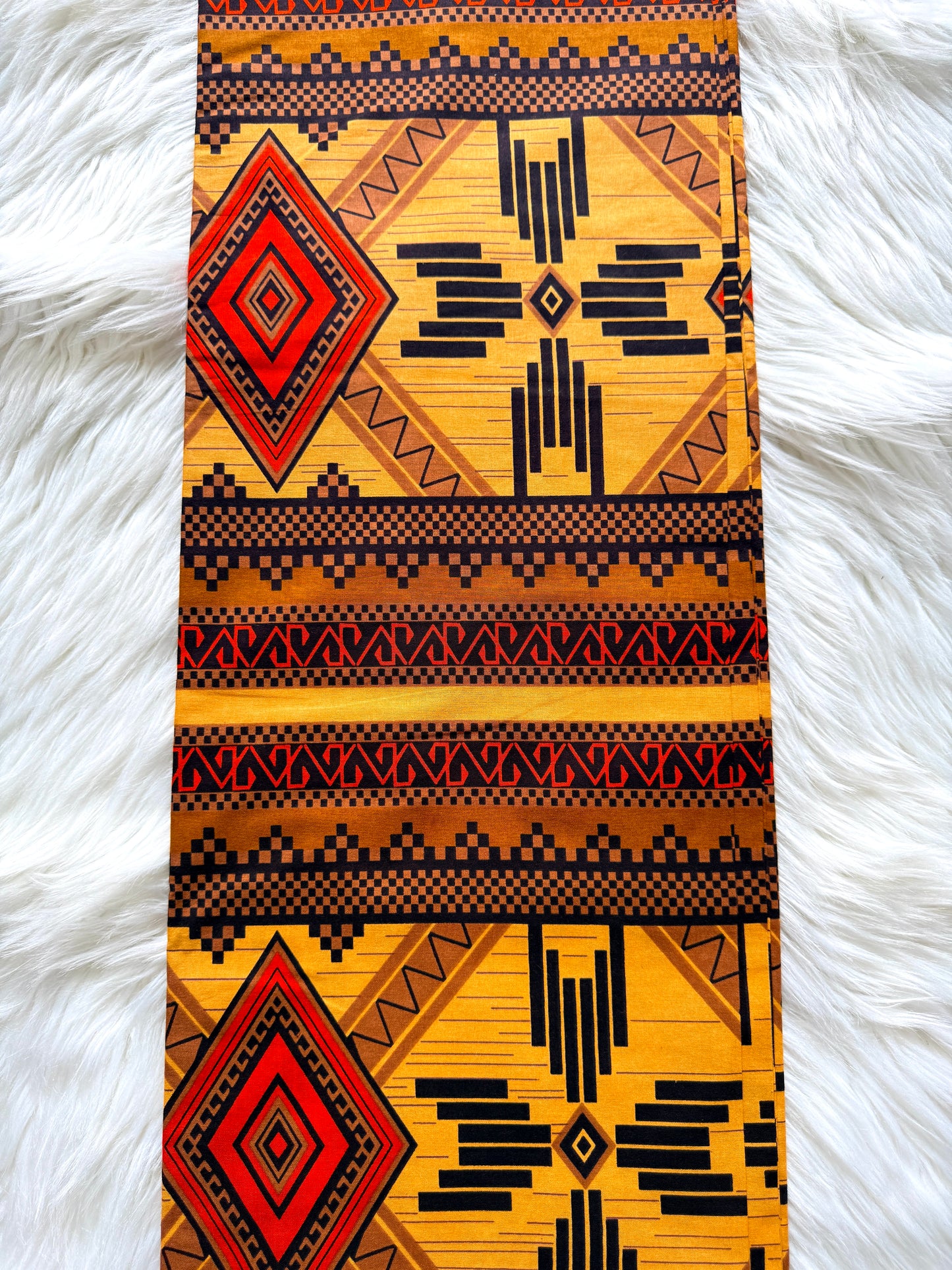 A long rectangular textile with a bold Southwestern geometric pattern in yellow, black, red, and brown, laid flat on a white faux fur surface.