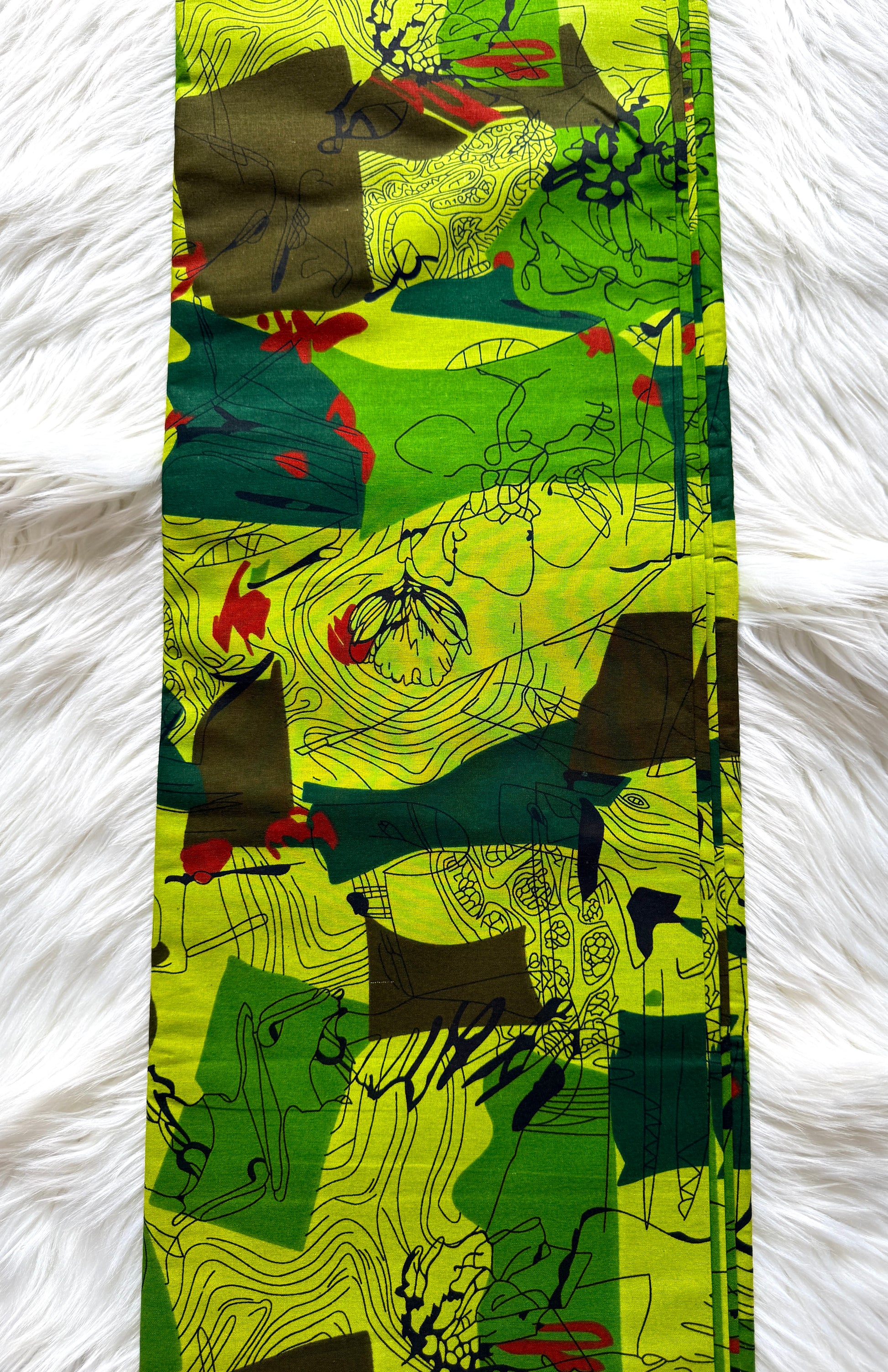 A long rectangular scarf laid flat on a white faux fur surface, featuring an abstract patchwork print with bright lime green, dark green, brown, black, and small red accents.
