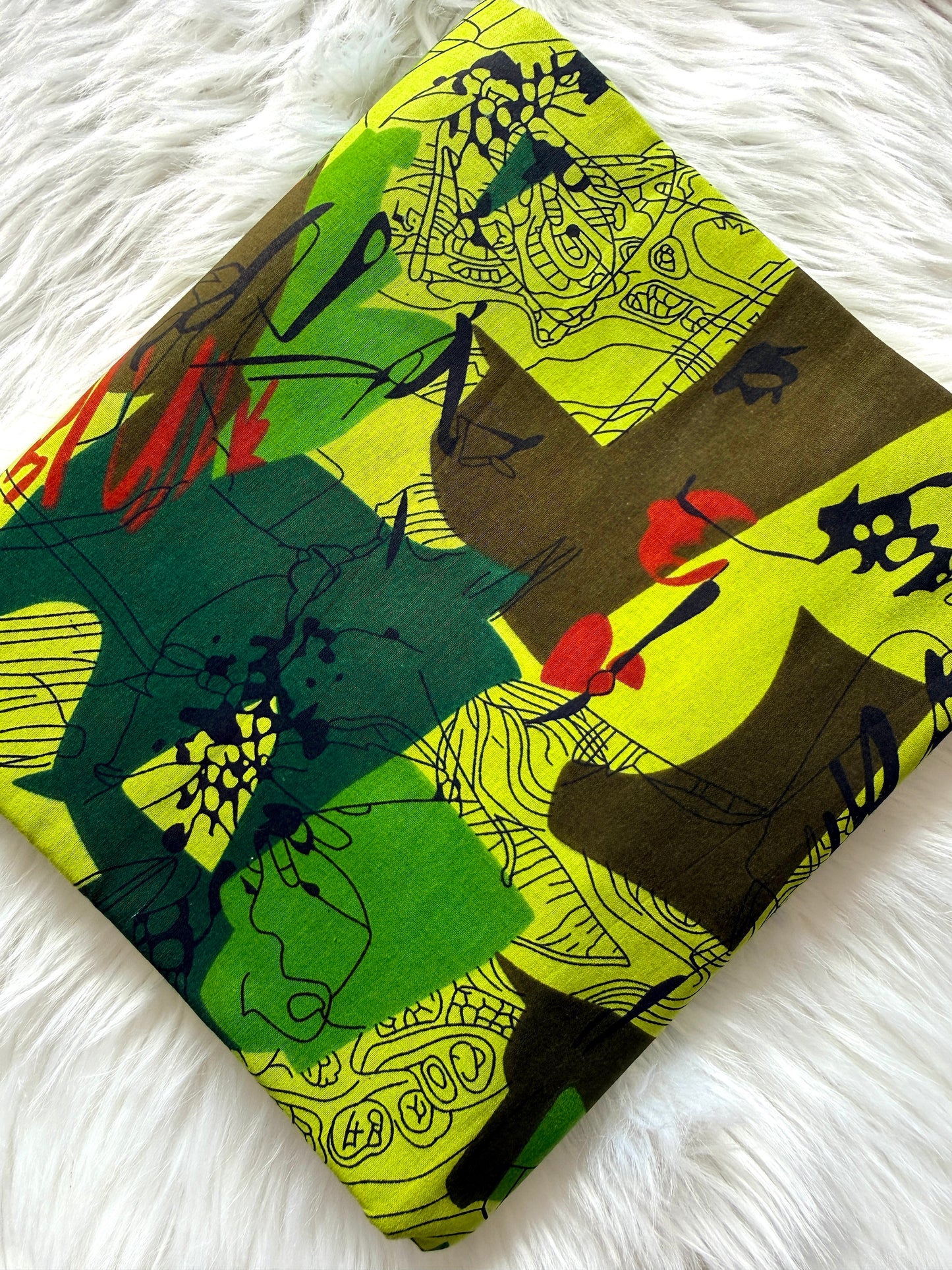 A folded square fabric item, likely a scarf or small bag, featuring an abstract multicolor print with lime green, forest green, brown, black, and red brushstrokes, displayed on a white faux fur surface.