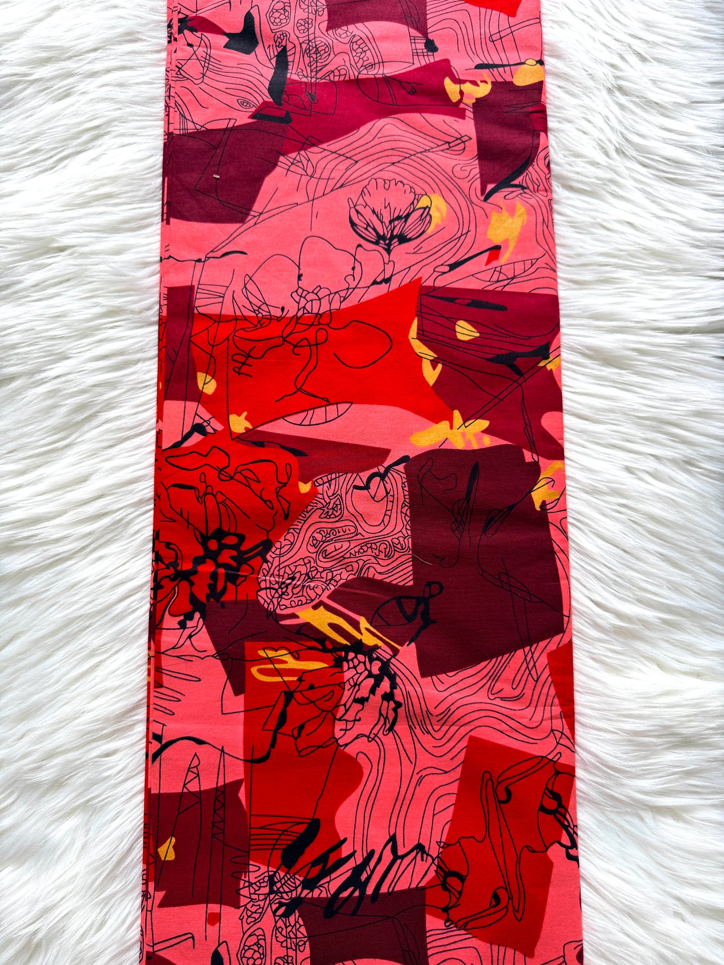 A long rectangular scarf laid flat on a white faux-fur surface. The scarf features an abstract, painterly pattern in shades of red, pink, and magenta with black line work and small yellow accents.