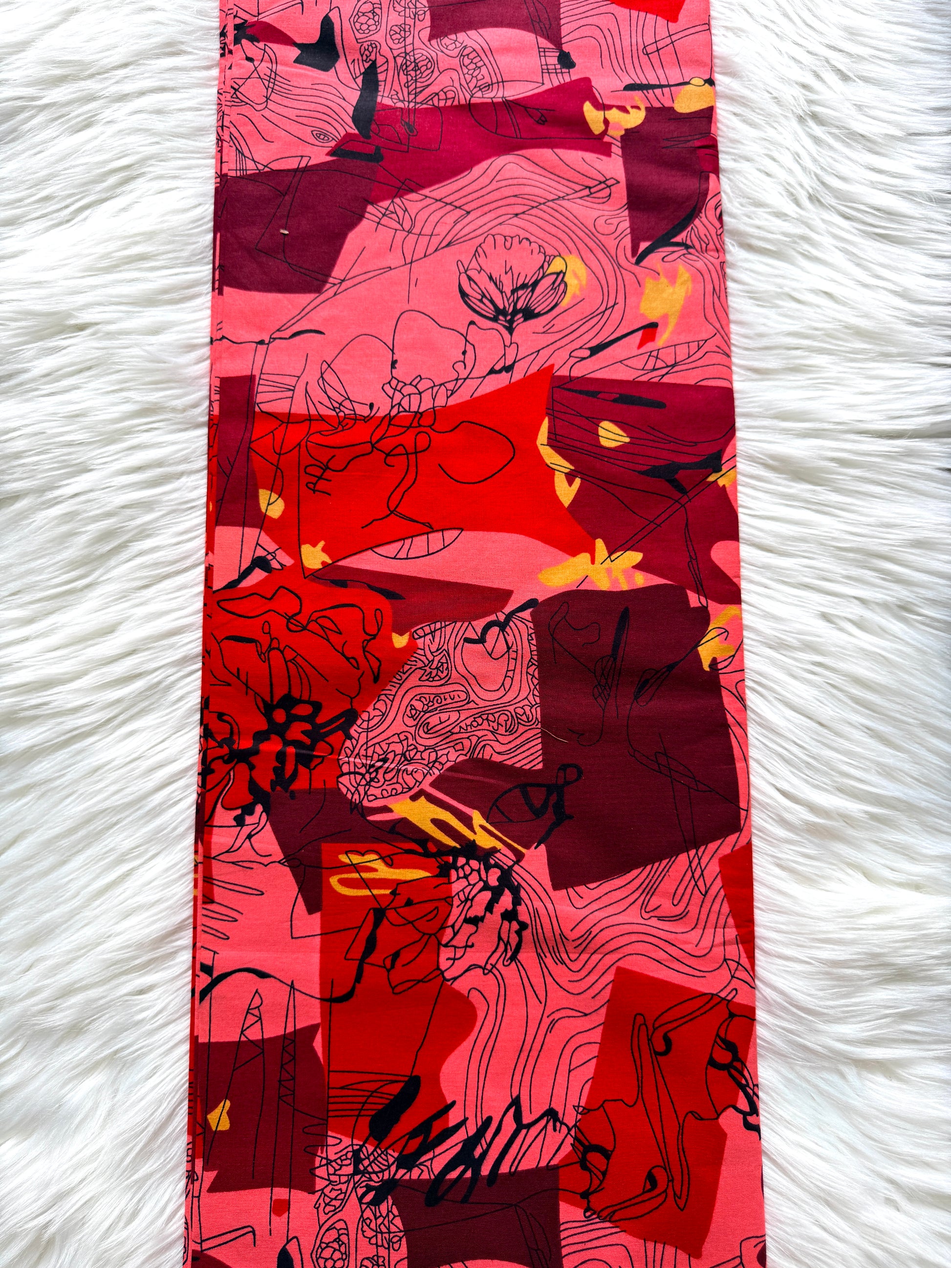 A long rectangular scarf laid flat on a white faux-fur surface. The scarf features an abstract, painterly pattern in shades of red, pink, and magenta with black line work and small yellow accents.