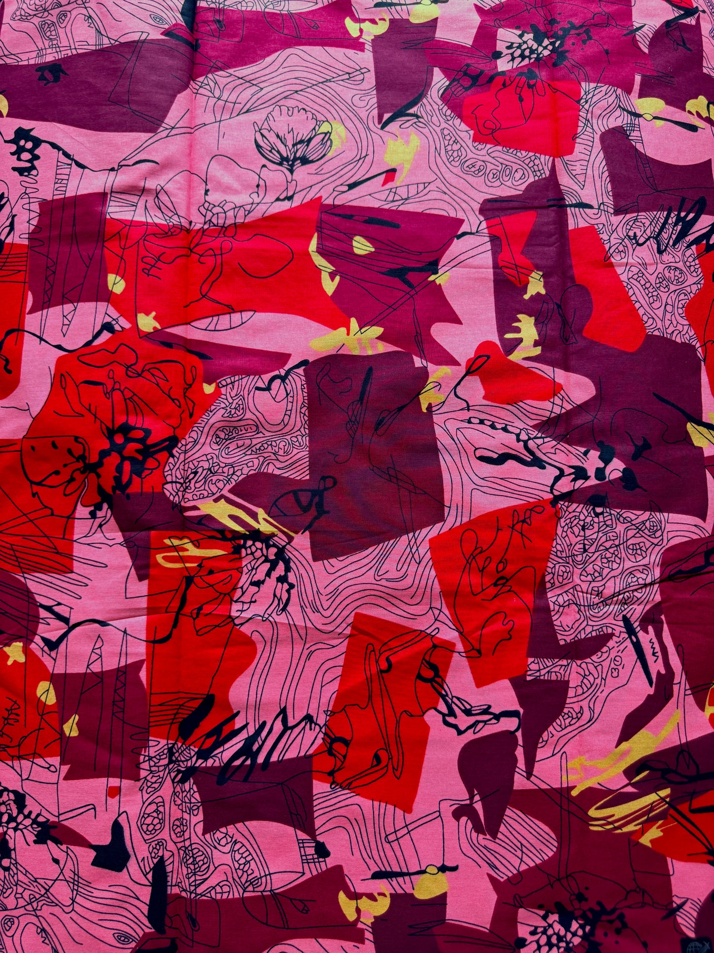 A close-up of a patterned fabric featuring an abstract design with overlapping pink, red, and deep purple blocks, overlaid with black sketch-like line art and small yellow accents.