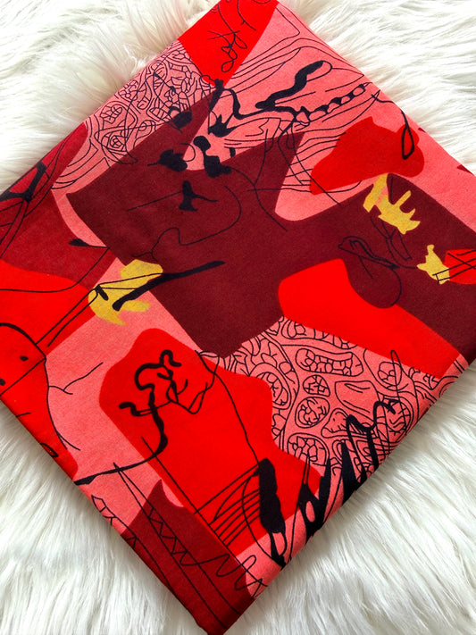 A folded square scarf featuring an abstract art print with red, pink, and dark maroon blocks, black sketch lines, and small yellow accents, displayed on a white faux fur surface.