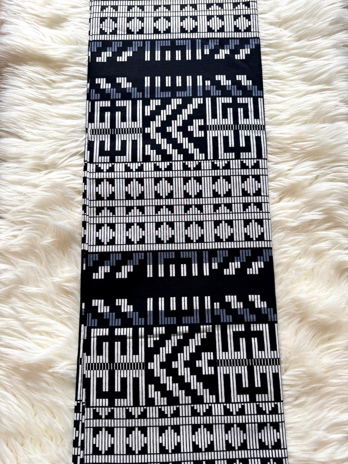 A long rectangular woven textile with a bold geometric pattern in black, white, and light blue, laid flat on a white faux fur surface.