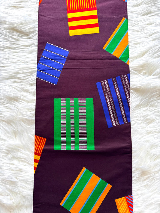 A dark purple fabric runner or table runner featuring a colorful geometric print with striped and patterned rectangular blocks in green, blue, yellow, orange, and red, laid flat on a white faux fur surface.