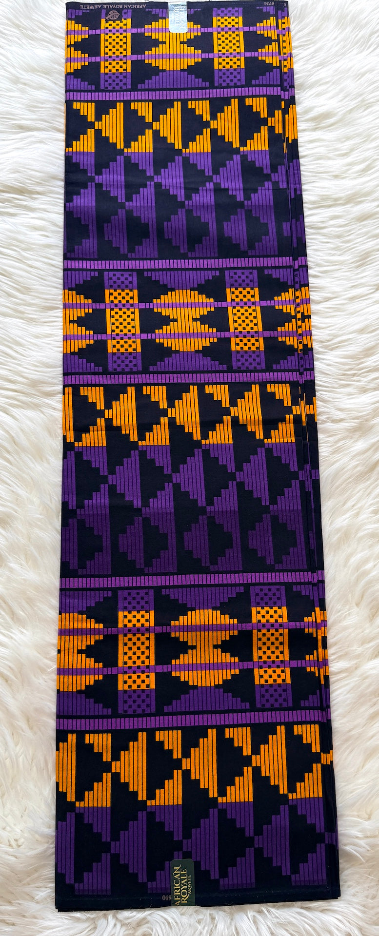 A long rectangular textile laid flat on a white faux-fur surface, featuring bold geometric and tribal-inspired patterns in purple, orange, and black.