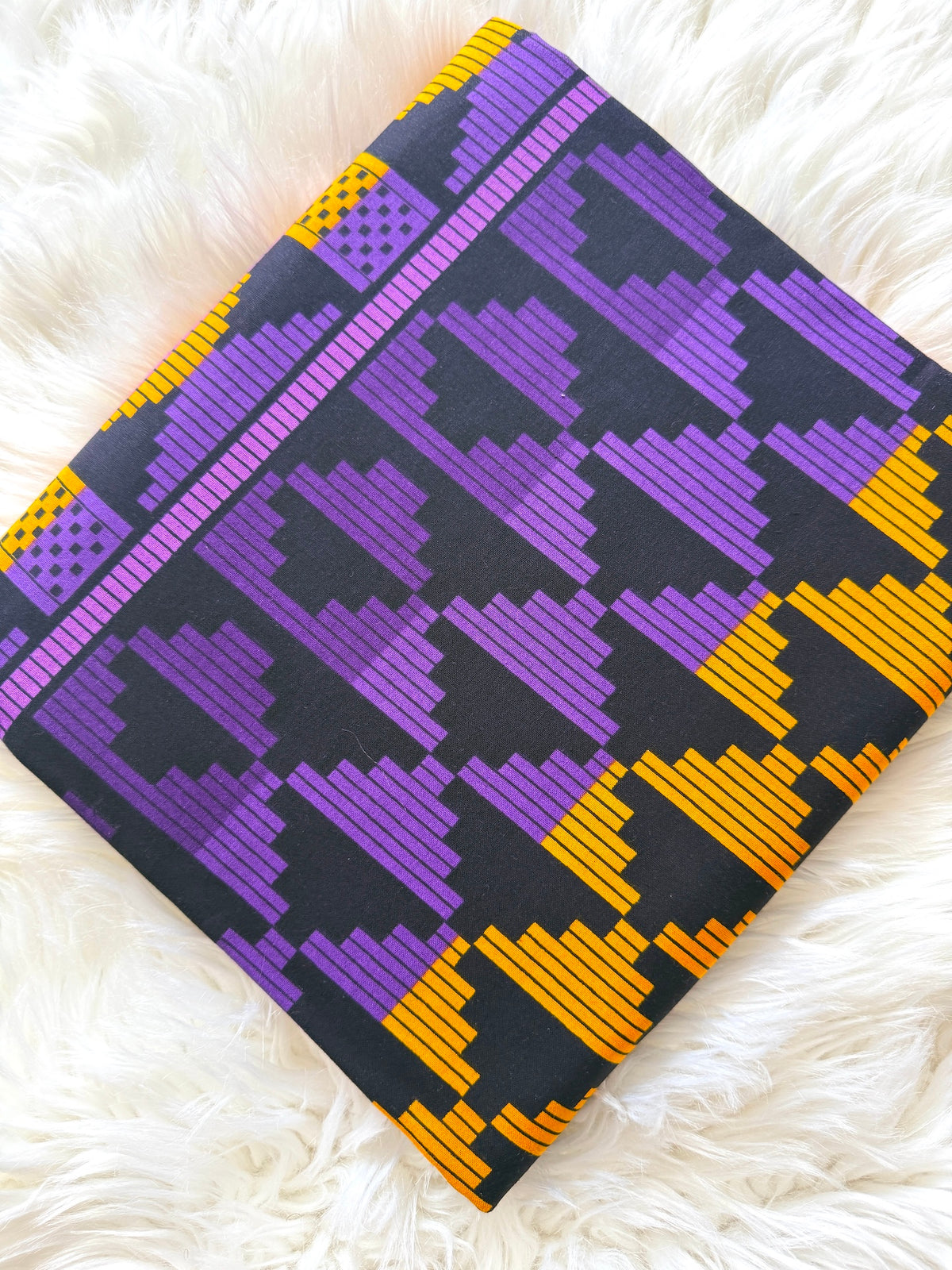 A folded textile product, likely a pillowcase or fabric panel, featuring a bold geometric pattern in purple, yellow, and black, displayed on a white faux fur background.