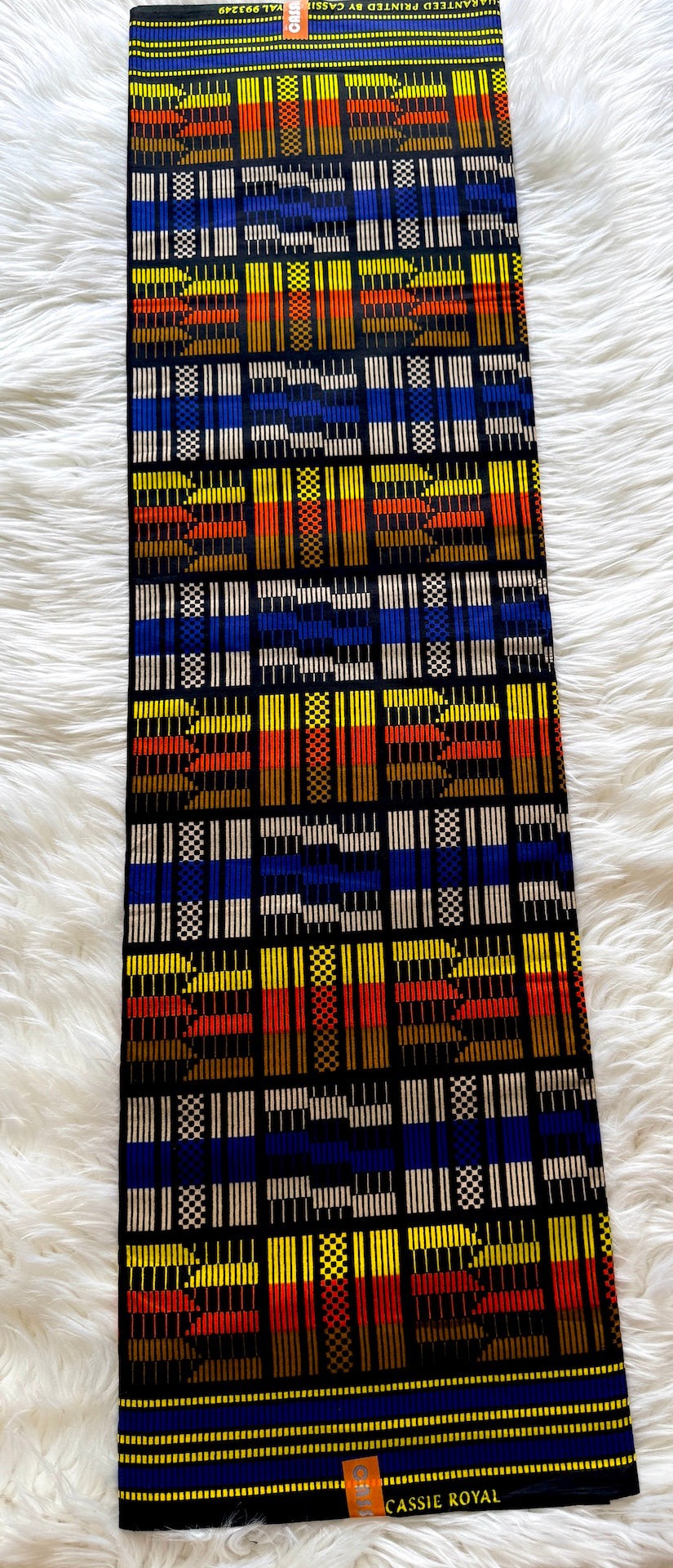 A long rectangular textile laid flat on a white faux-fur surface, featuring a bold multicolor geometric pattern with black, blue, red, yellow, orange, and white blocks. The brand name 'CASSE ROYAL' is visible on small orange labels near the top and bottom edges.