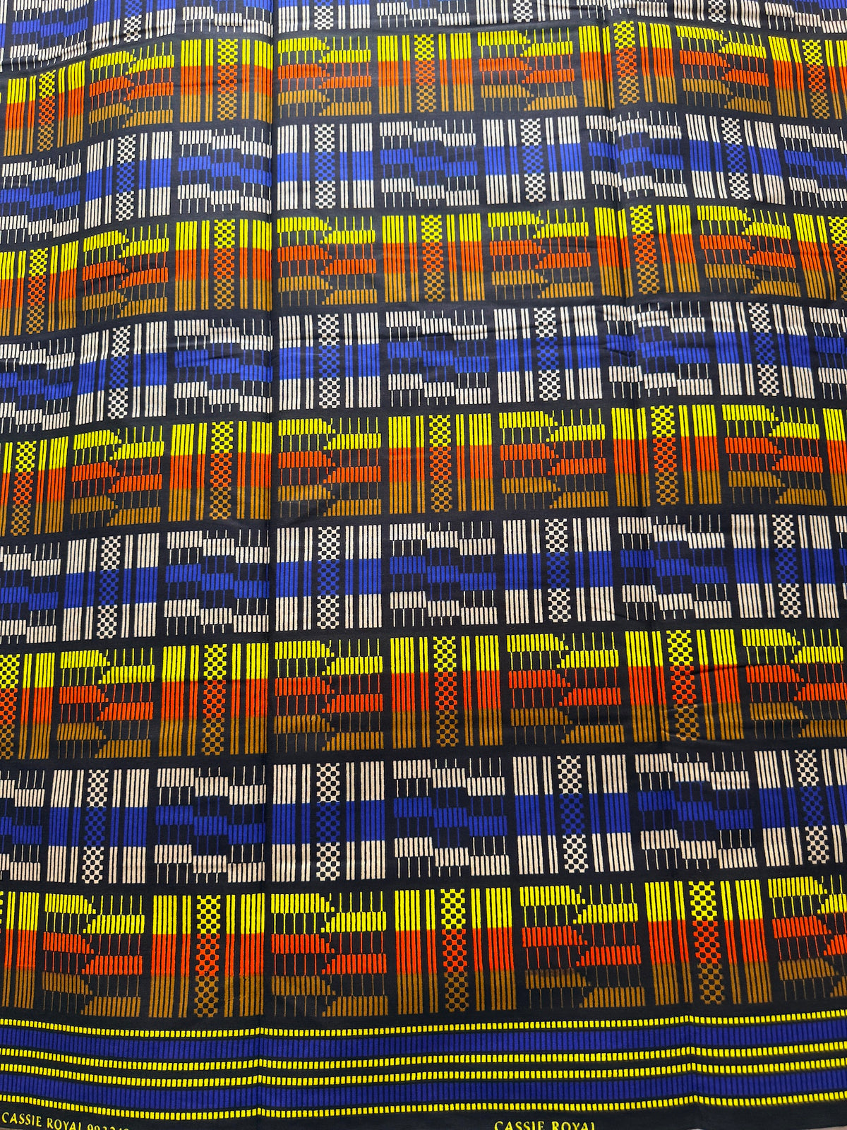 A close-up of a woven fabric with a bold geometric pattern in blue, yellow, orange, red, and white on a black background. The fabric has a decorative border at the bottom with the text 'CASSE ROYALE' in yellow.