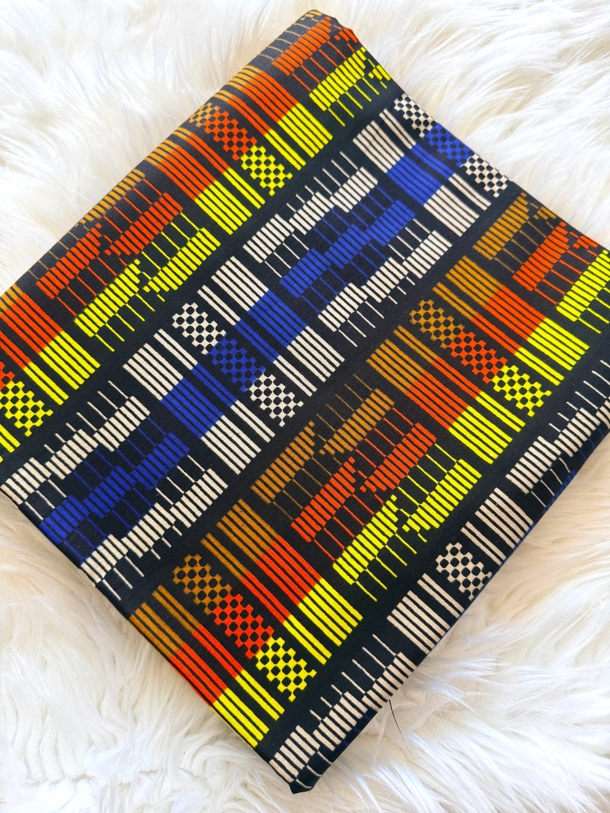 A square woven textile or pillow cover with a bold geometric pattern in black, yellow, blue, orange, and white, laid flat on a white faux fur surface.