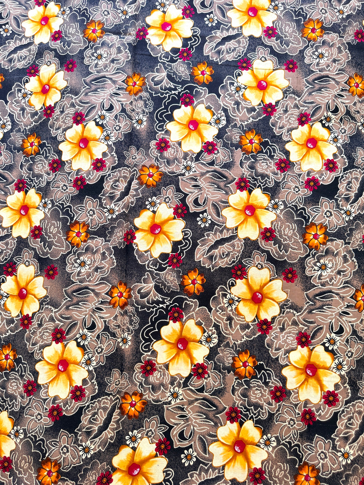 A close-up of a patterned fabric featuring large yellow and orange flowers with red centers, layered over a gray and white floral lace-like overlay on a dark background.