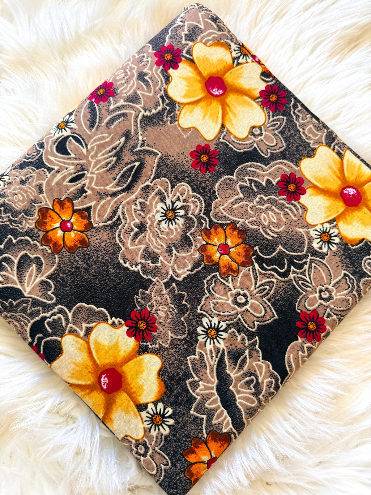 A square fabric item, likely a pillow cover or scarf, featuring a retro floral pattern with yellow, orange, and red flowers on a dark brown and beige background, displayed on a white faux fur surface.
