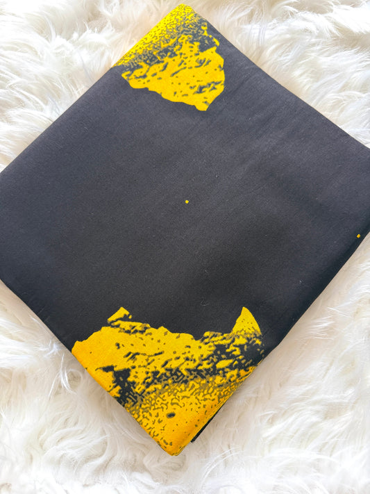 A square black fabric item, likely a pouch or small bag, featuring two distressed yellow graphic accents near the top and bottom corners, displayed on a white faux fur background.