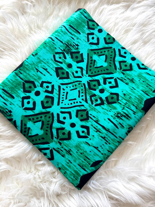 A square fabric item, likely a pillow cover or small blanket, featuring a vibrant turquoise and teal pattern with black geometric tribal-style motifs, laid flat on a white faux fur surface.