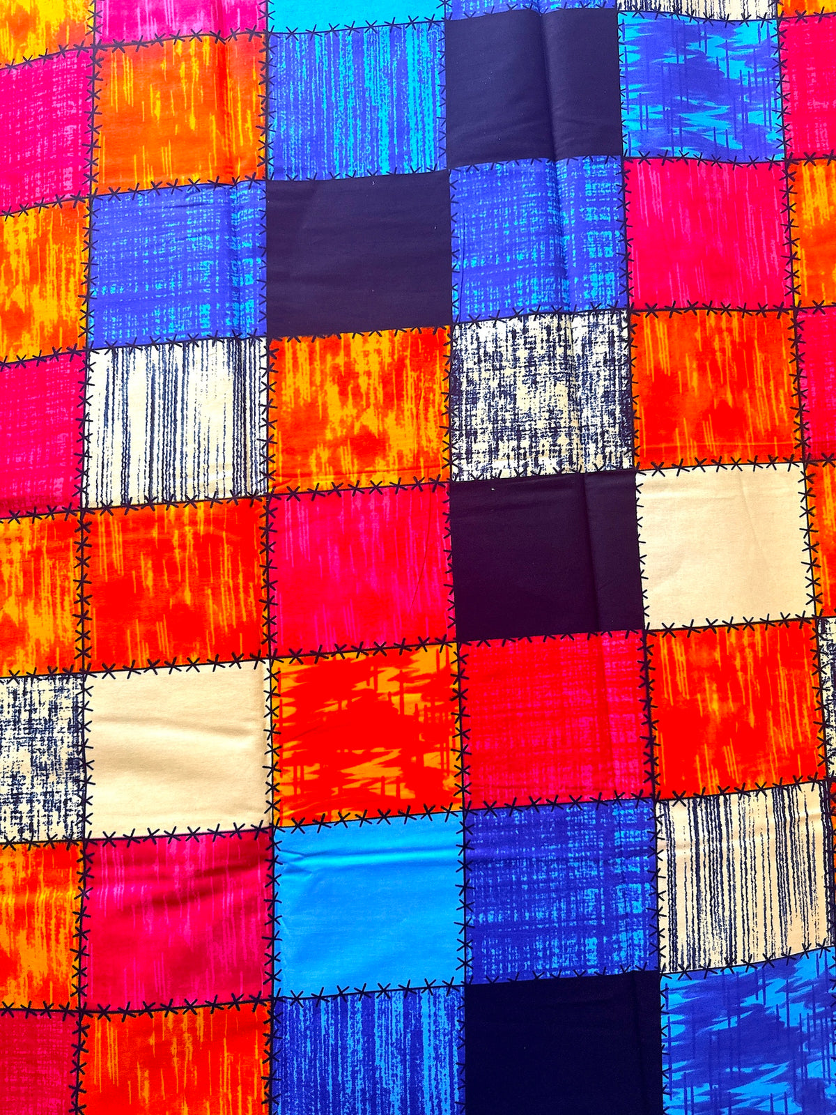 A close-up of a colorful patchwork fabric with stitched square panels in bright blue, red, orange, pink, cream, and dark navy, showing varied textures.