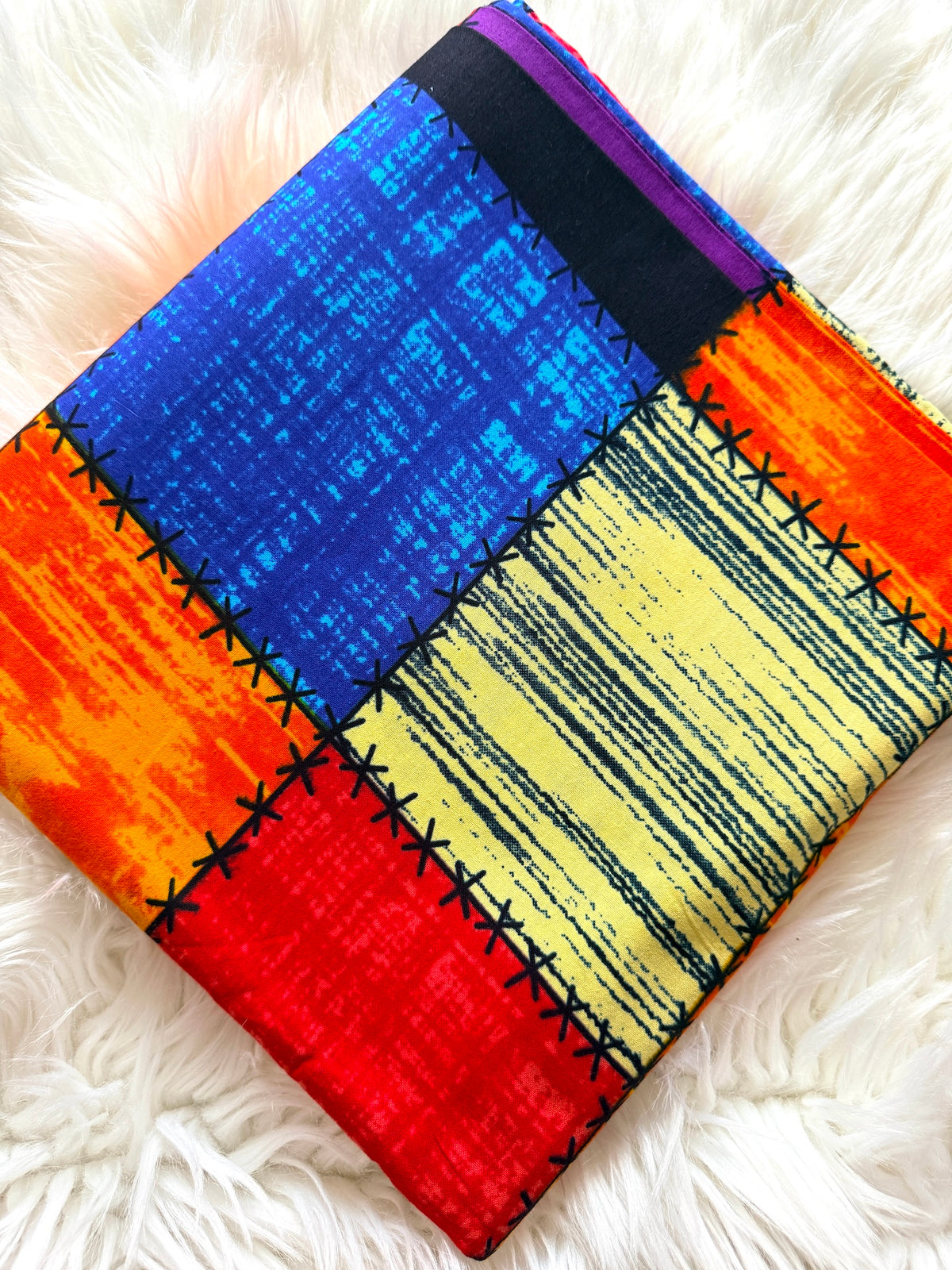 A square patchwork fabric pouch or small bag laid flat on a white faux-fur surface. The fabric has large stitched color blocks in blue, yellow, orange, red, black, and purple, with visible black stitched seams.
