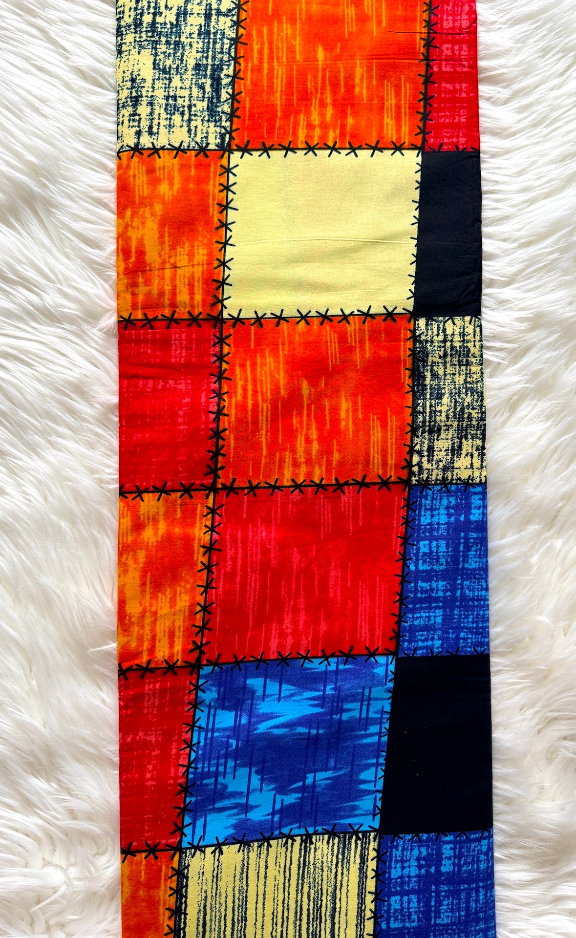A long rectangular patchwork textile laid flat on a white faux-fur surface. The fabric features stitched square panels in orange, red, yellow, blue, black, and cream, with textured and striped patterns.