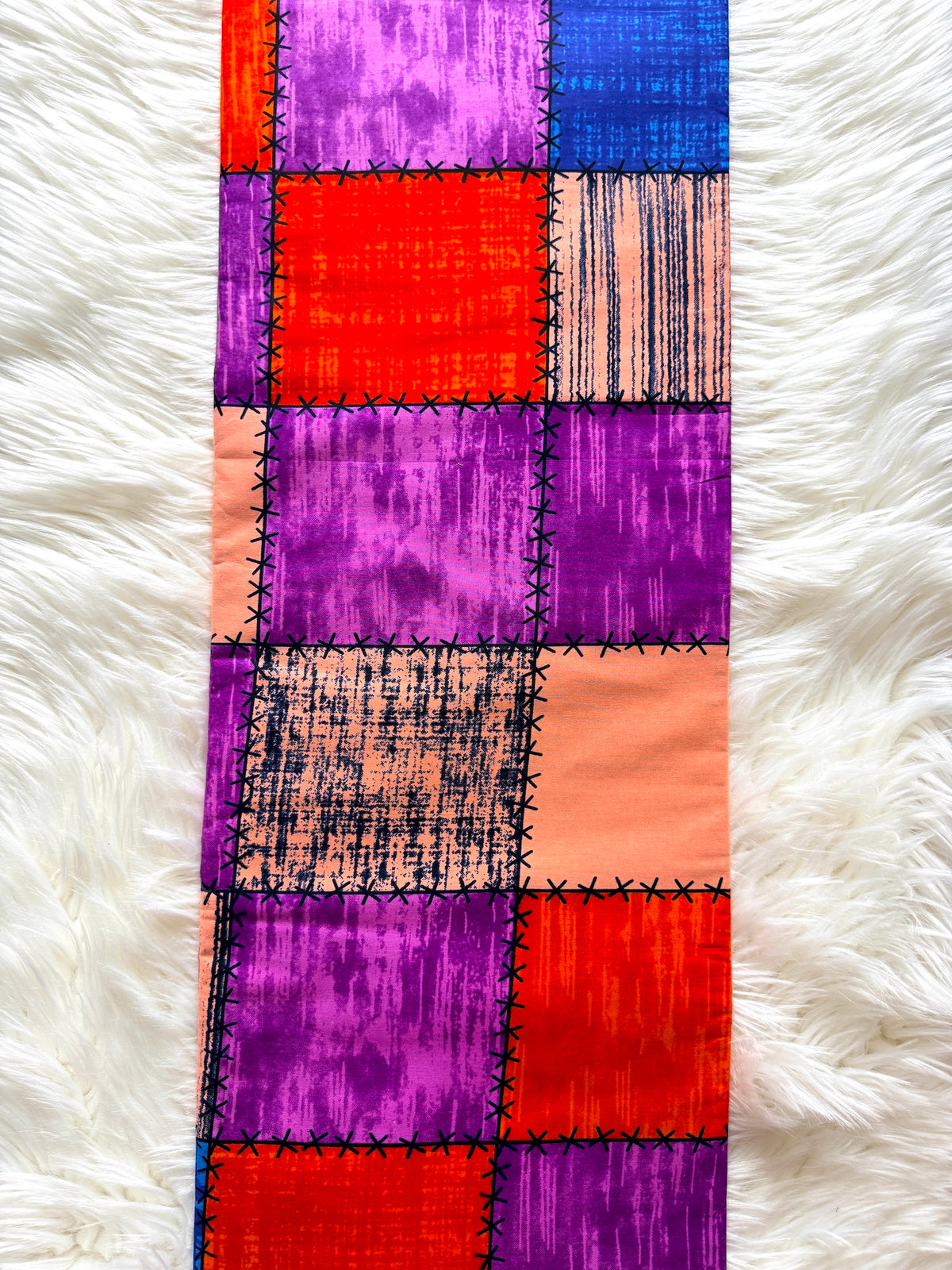 A long rectangular textile runner laid flat on a white faux-fur surface. The runner has a patchwork design made of stitched square panels in bright purple, coral pink, orange-red, blue, and peach tones with black stitched seams.