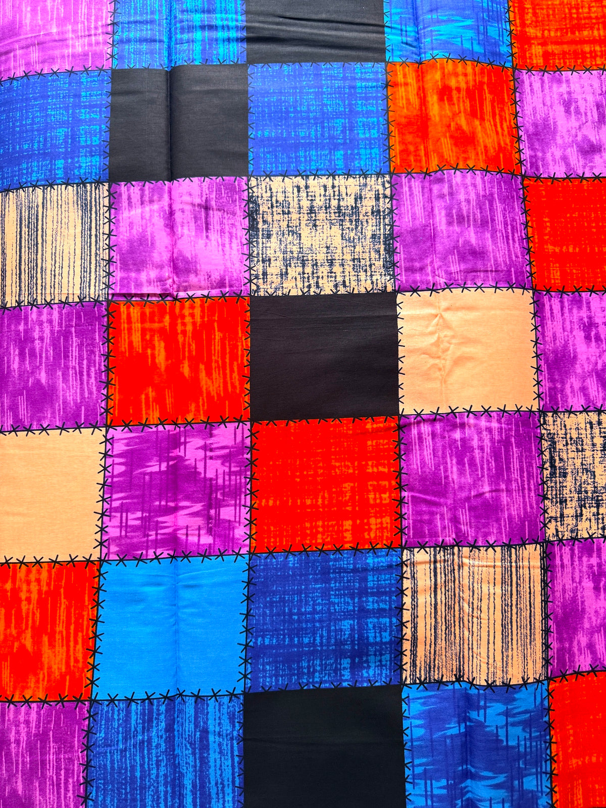 A close-up of a colorful patchwork fabric with stitched square panels in bright blue, purple, orange, red, black, and beige tones.