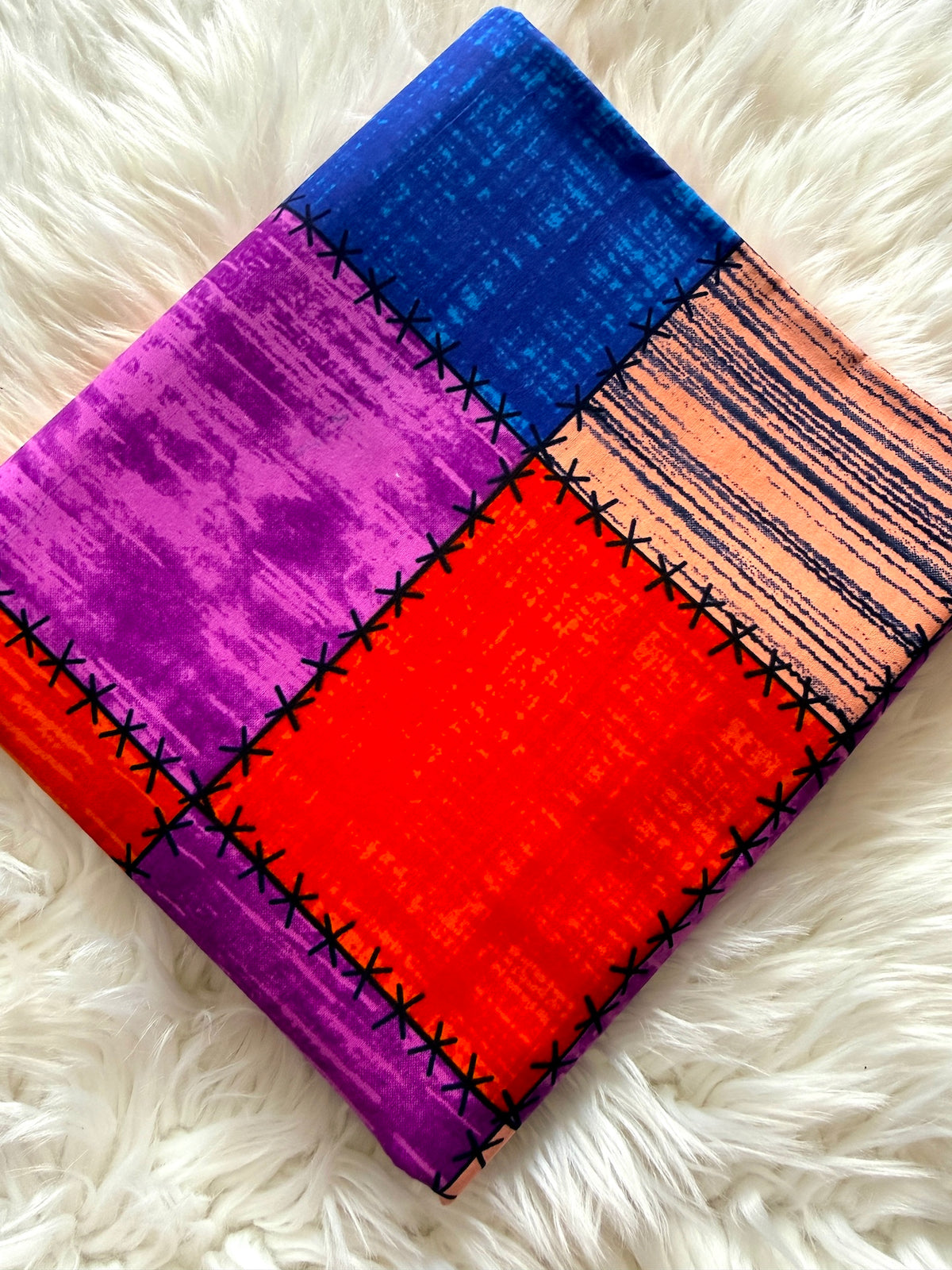 A folded square textile with a patchwork design in blue, purple, orange, and red, featuring black stitched seam details, displayed on a white faux fur background.