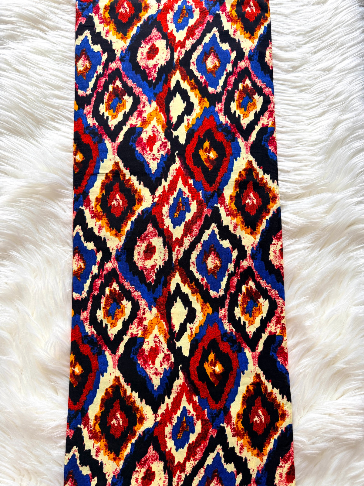 A long rectangular textile laid flat on a white faux-fur surface, featuring a bold repeating diamond ikat-style pattern in red, blue, black, cream, and orange.