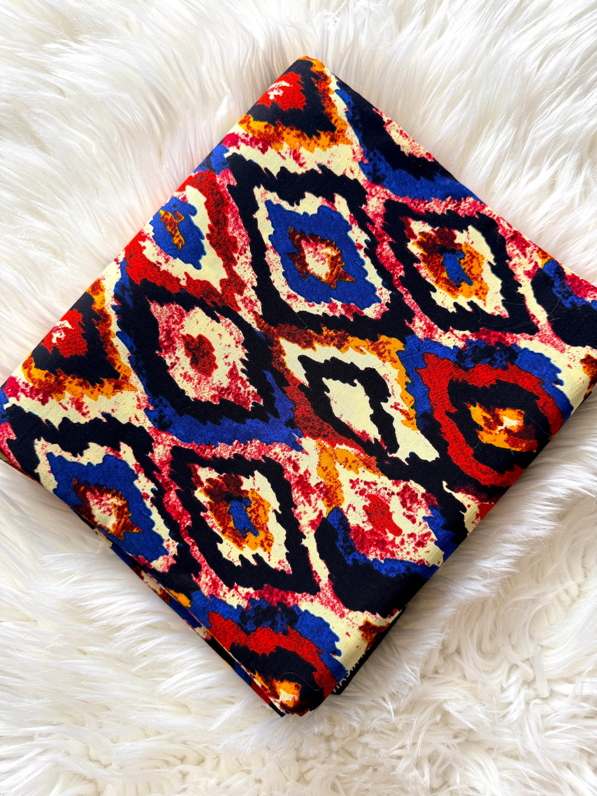 A square fabric item, likely a scarf or small bag, featuring a vibrant, multicolor ikat diamond pattern, displayed diagonally on a white faux fur surface.