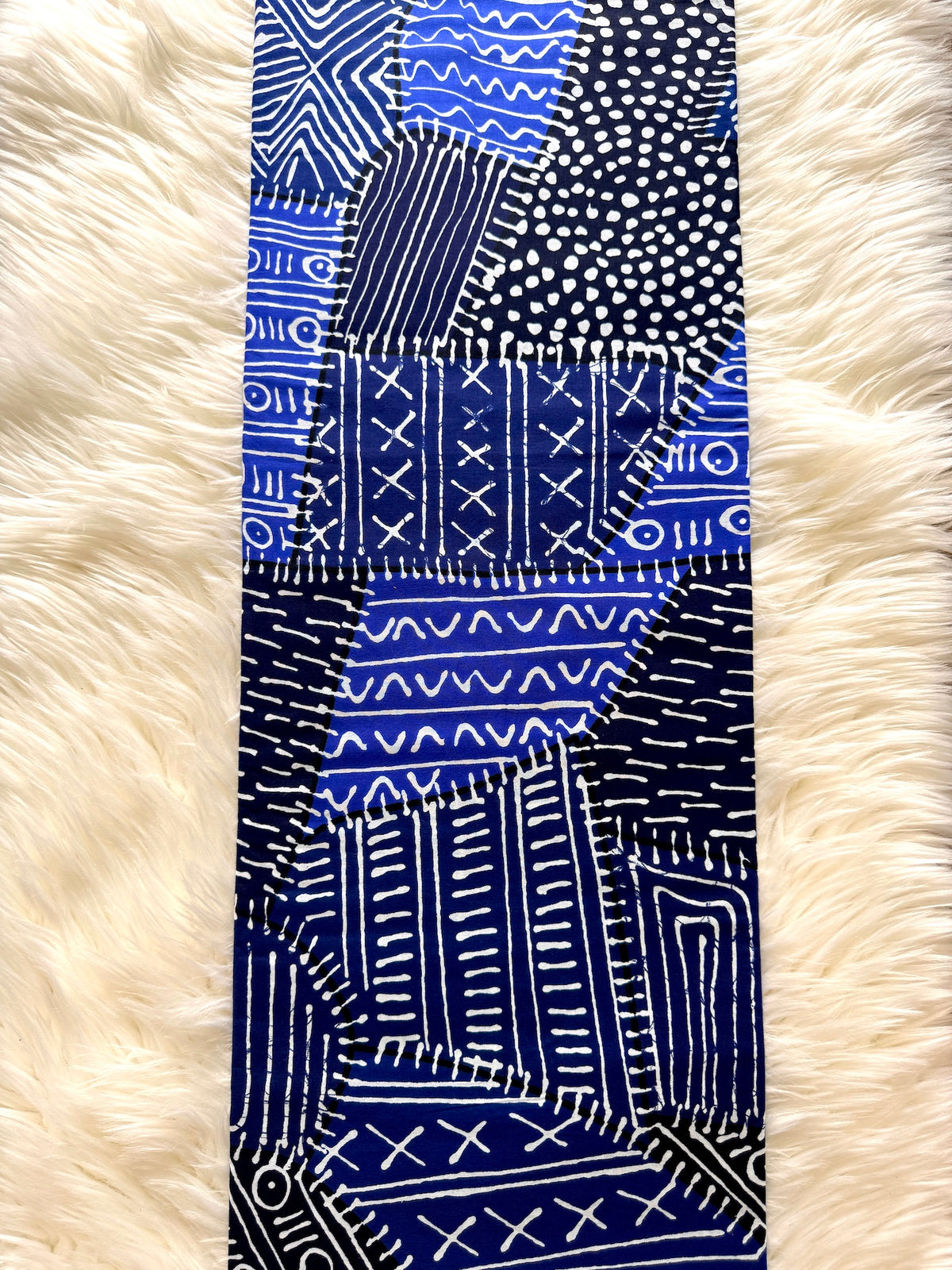 A vertical textile panel or runner featuring a patchwork of blue, navy, black, and white hand-drawn tribal patterns, laid flat on a cream faux fur background.