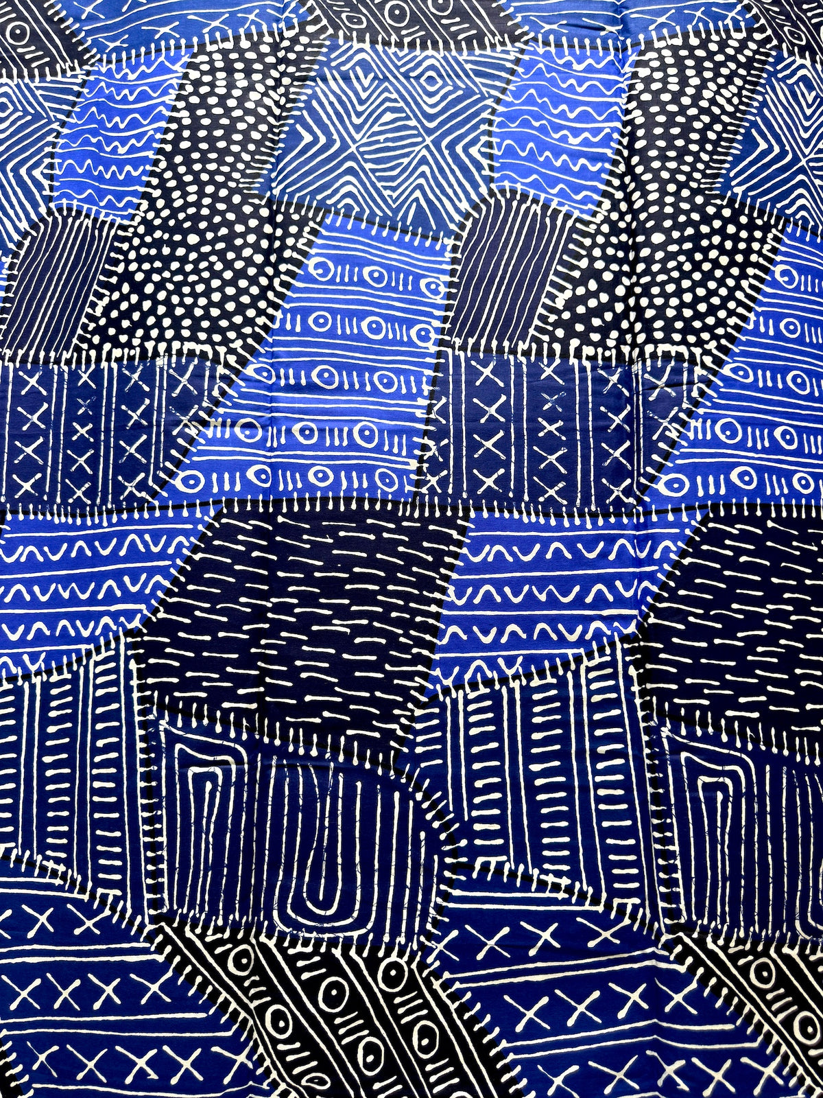 A close-up of a blue and black patterned fabric with white hand-drawn geometric and dotted designs arranged in a patchwork layout.