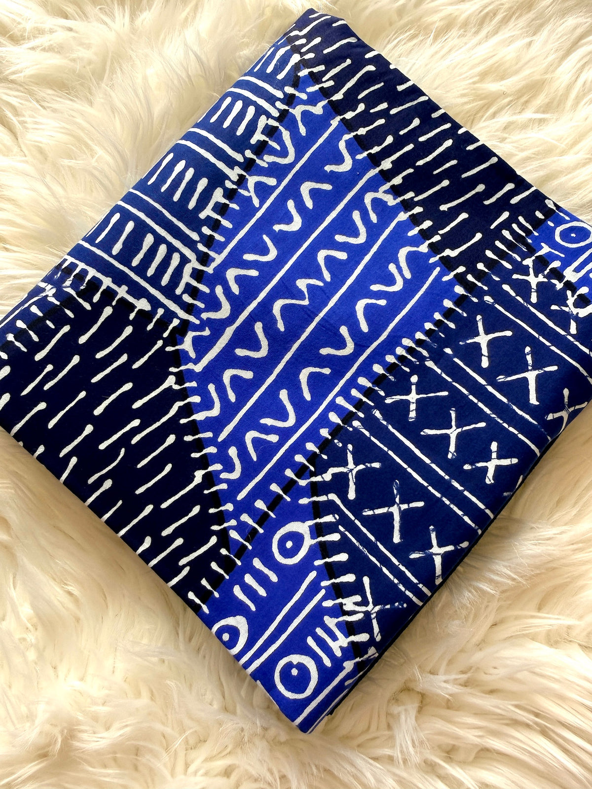 A square fabric item, likely a small pouch or clutch, featuring a vibrant blue and navy indigo batik pattern with white hand-drawn tribal motifs, displayed on a soft white faux fur surface.